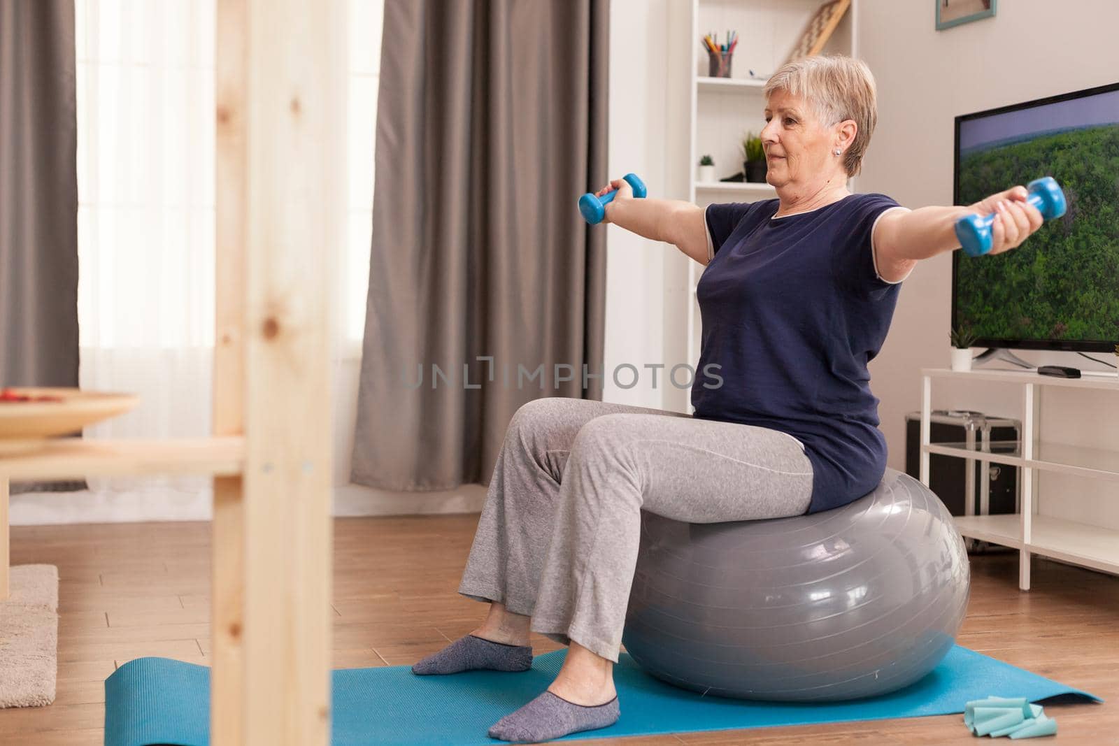 Retired woman doing sports for health in living room. Old person pensioner online internet exercise training at home sport activity with dumbbell, resistance band