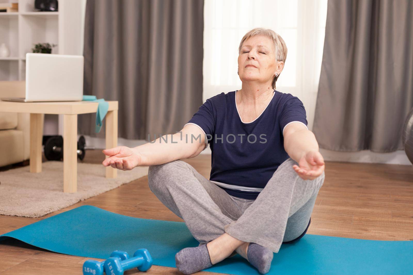 Peaceful senior woman with eyes closed doing yoga in living room. Old person pensioner online internet exercise training at home sport activity with dumbbell, resistance band, swiss ball at elderly retirement age