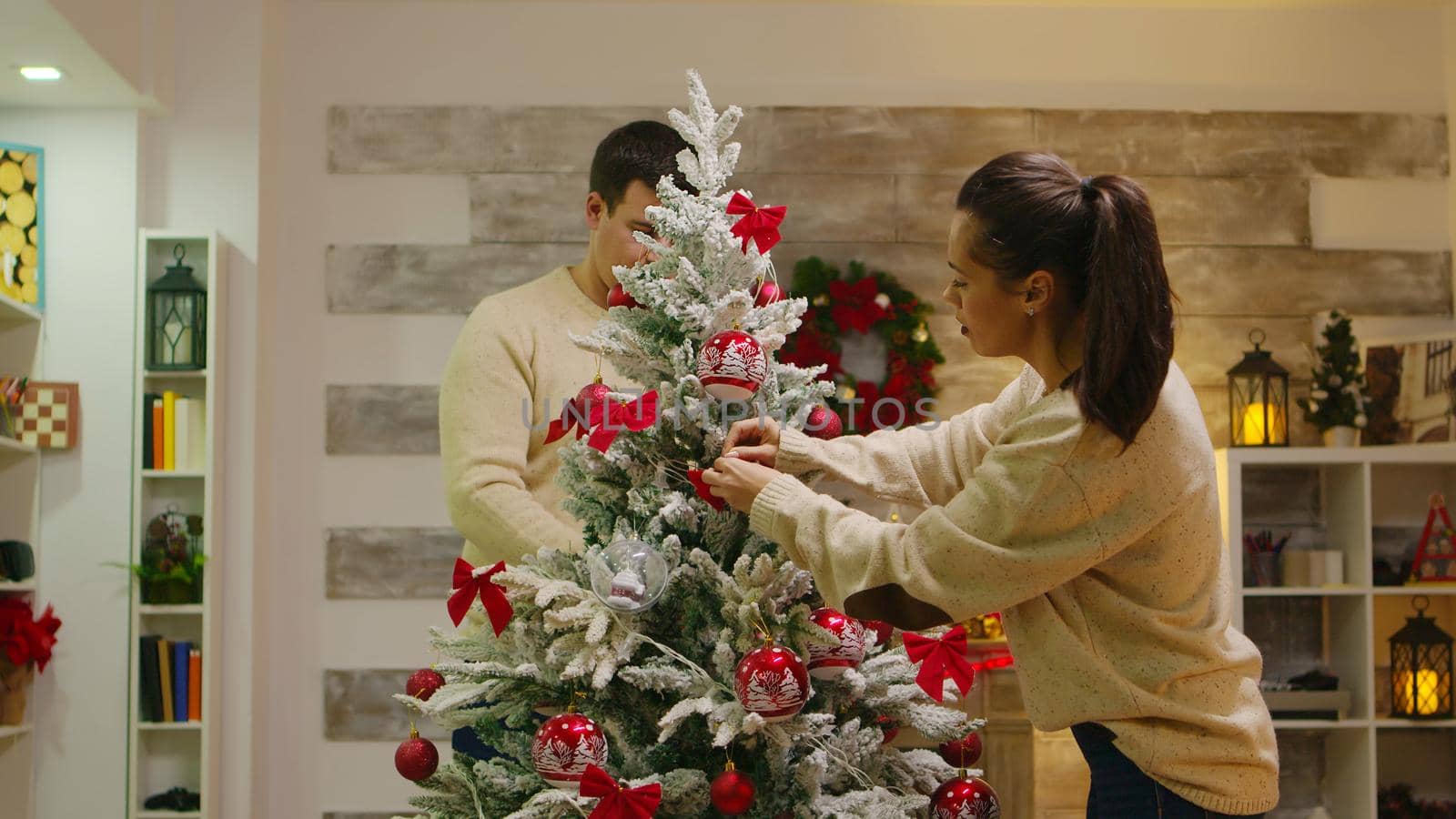 Young couple in love assembled christmas tree in living room. Romantic couple. Decorating beautiful xmas tree with glass ball decorations. Wife and husband in matching clothes helping ornate home with garland lights