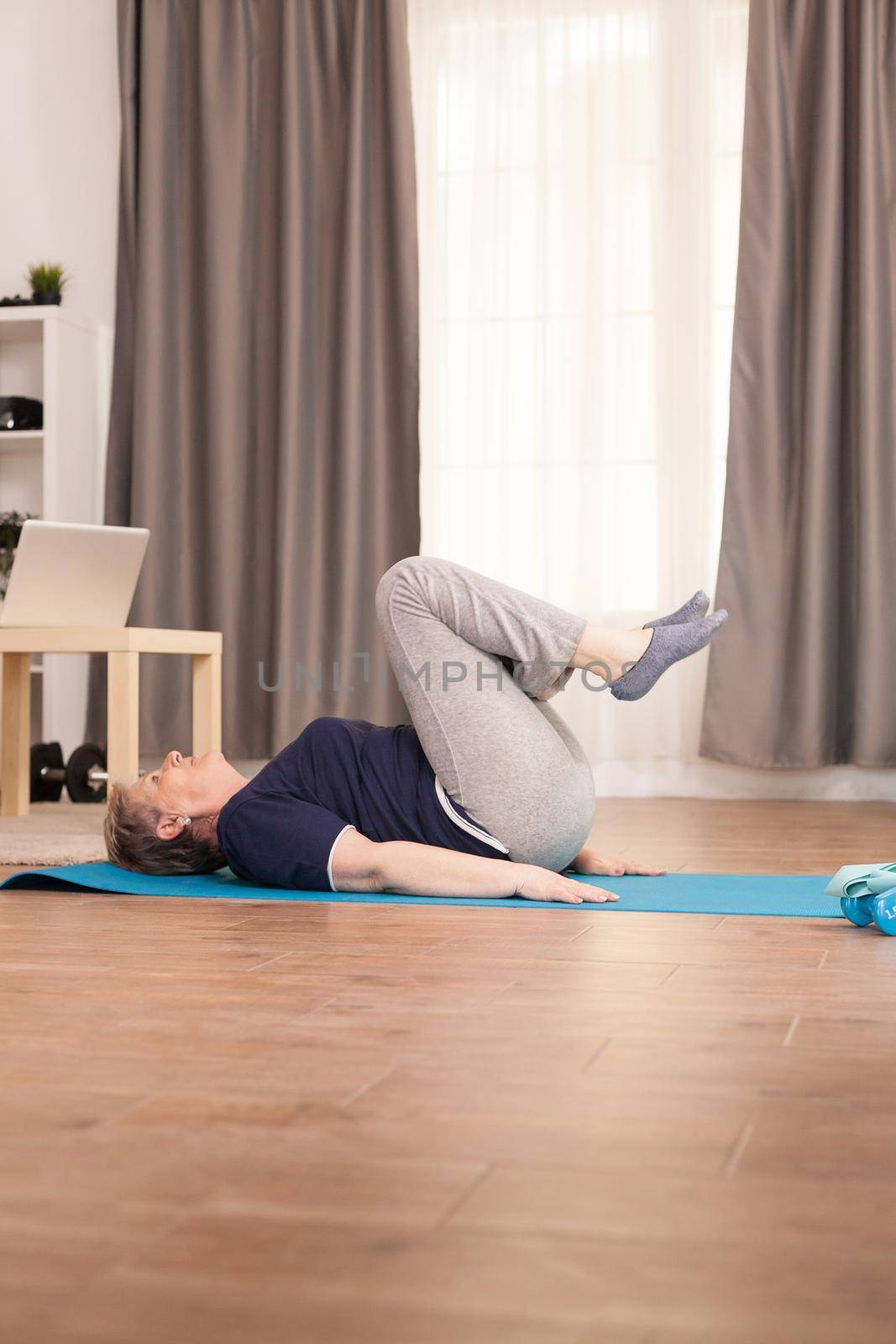 Elderly age woman doing abs physical training in living room on the yoga mat. Old person pensioner online internet exercise training at home sport activity with dumbbell, resistance band, swiss ball at elderly retirement age