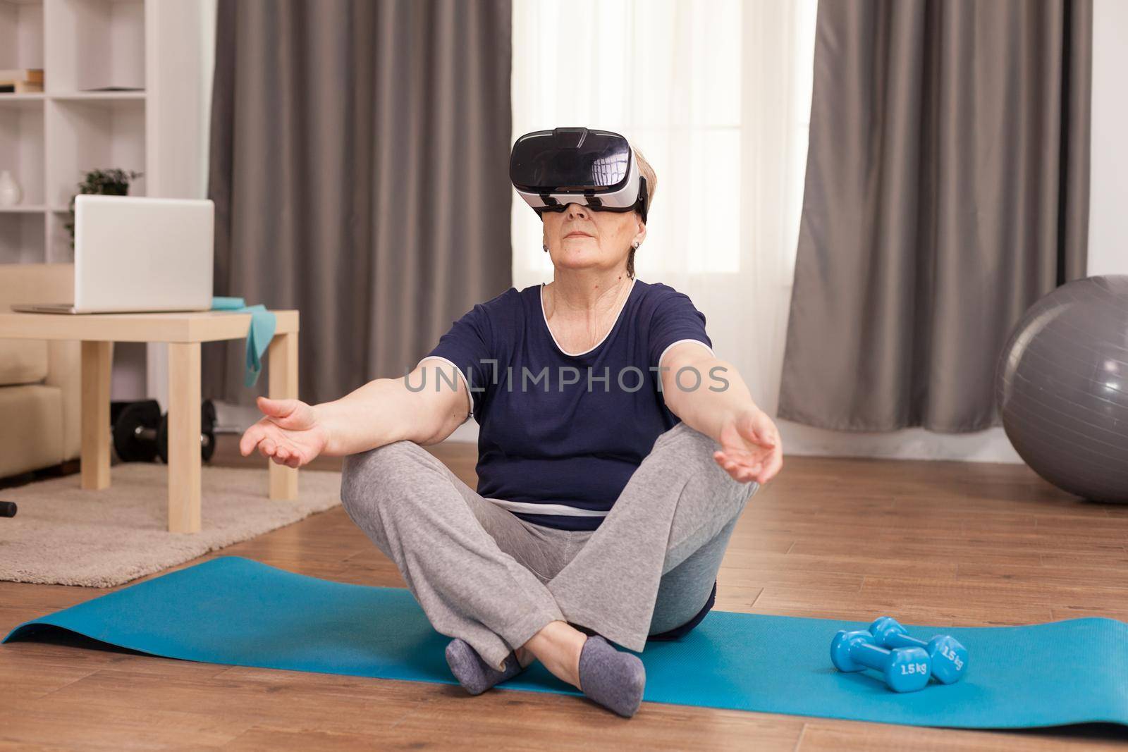 Grandmother wearing VR headset sitting on yoga mat. Old person pensioner online internet exercise training at home sport activity with dumbbell, resistance band, swiss ball and VR headset at elderly retirement age