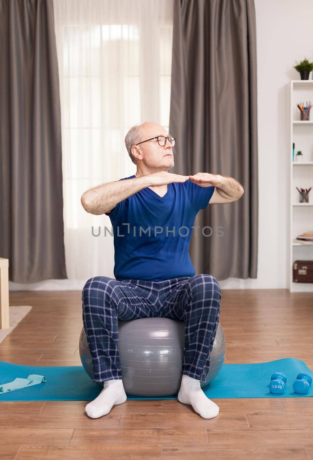 Retired man with healthy lifestyle doing arms exercises. Old person pensioner online internet exercise training at home sport activity with dumbbell, resistance band, swiss ball at elderly retirement age.