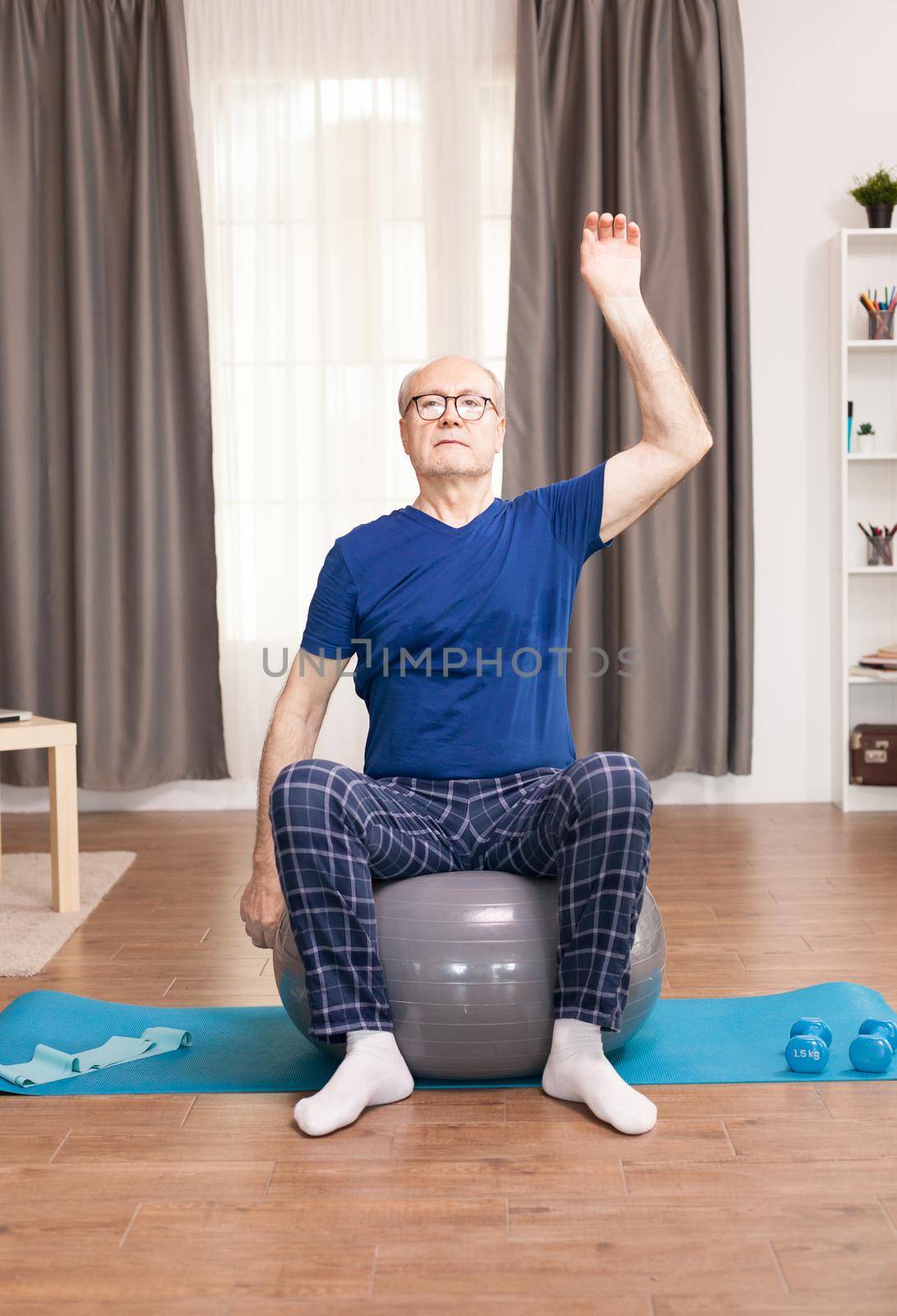 Old man with a healty lifestyle training on living room. Old man training on living room Old person pensioner online internet exercise training at home sport activity with dumbbell, resistance band, swiss ball at elderly retirement age.