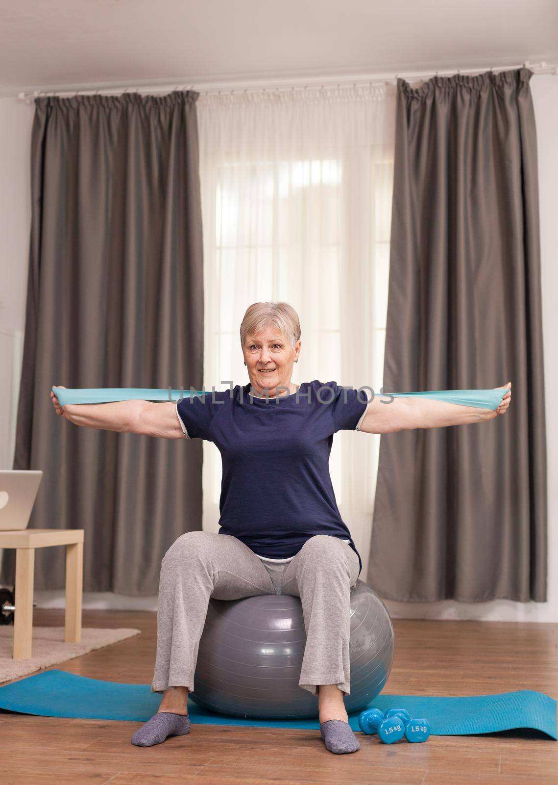 Retired woman with a healthy mind, sitting on a balance ball and training with an elastic band. Old person pensioner online internet exercise training at home sport activity with dumbbell, resistance band, swiss ball at elderly retirement age