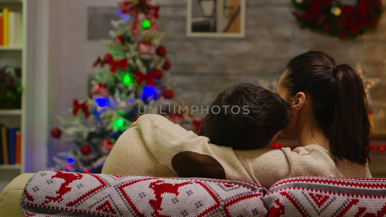 Back view of romantic couple sitting on couch on christmas day with xmas tree in the background. Xmas spirit for caucasian wife and husband celebrating positive happy new year eve sitting whispering merry christmas