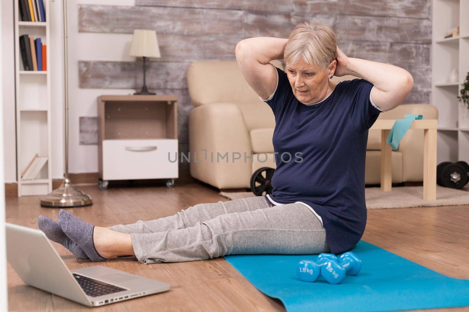 Aged woman doing online training sitting on yoga mat at home. Old person pensioner online internet exercise training at home sport activity with dumbbell, resistance band, swiss ball at elderly retirement age