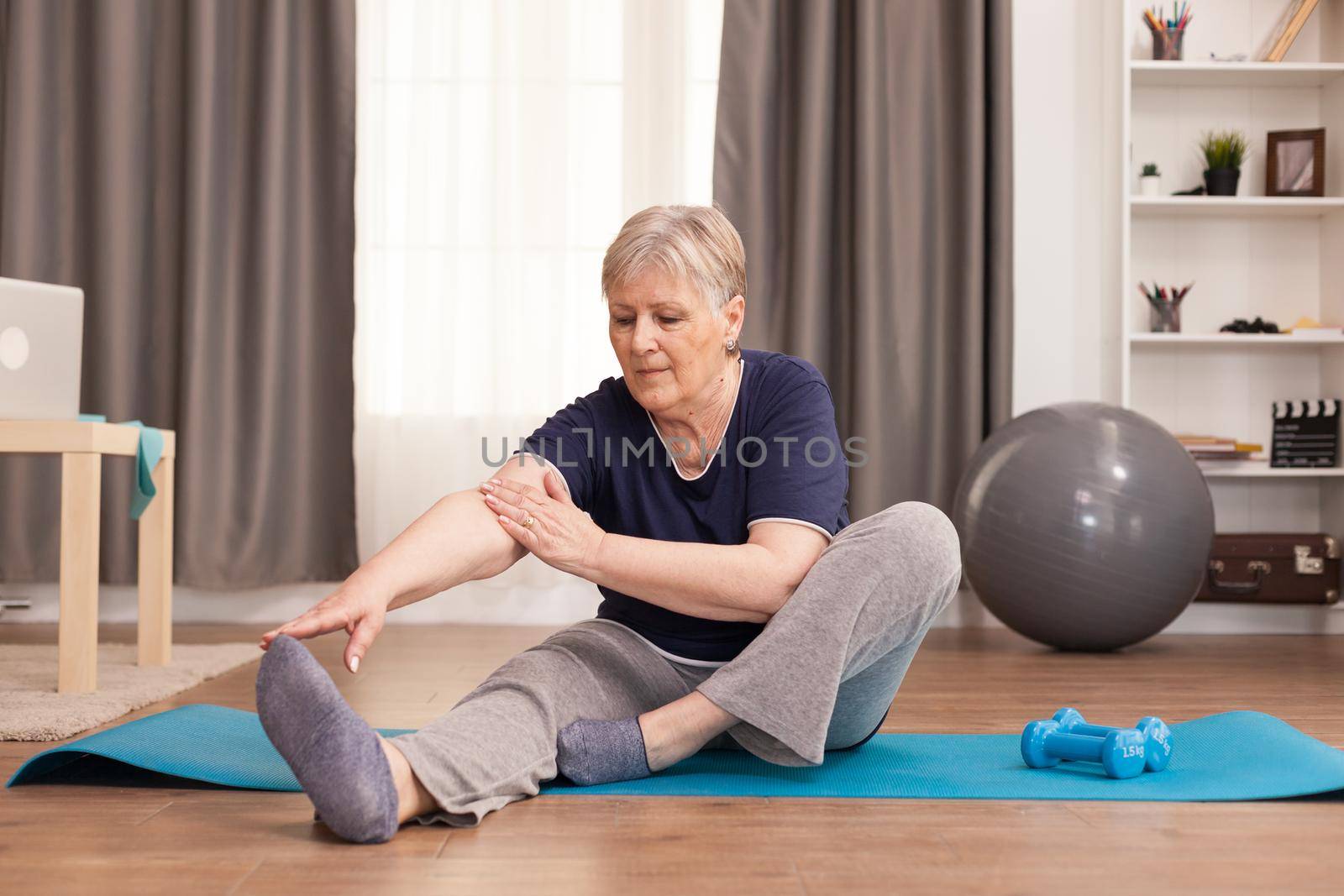 Active old woman exercising on the yoga mat in her comfortable apartment. Old person pensioner online internet exercise training at home sport activity with dumbbell, resistance band, swiss ball at elderly retirement age