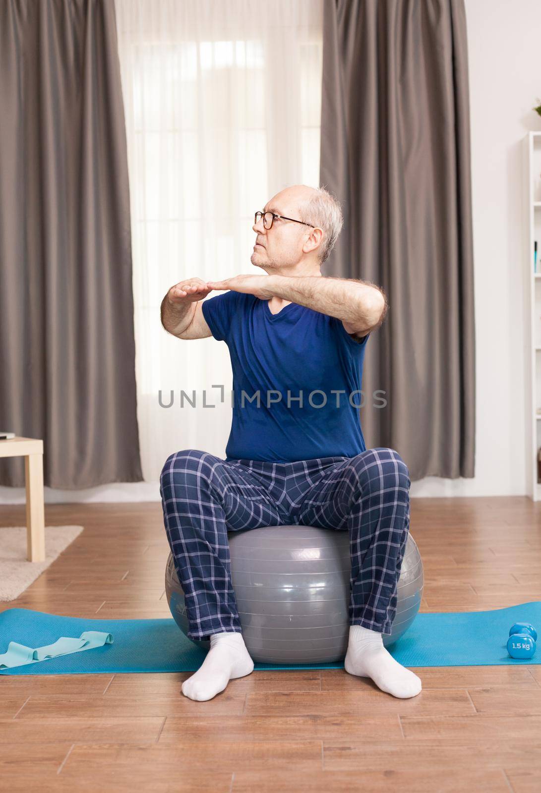 Grandfather training on swiss ball in living room. Old person pensioner online internet exercise training at home sport activity with dumbbell, resistance band, swiss ball at elderly retirement age.