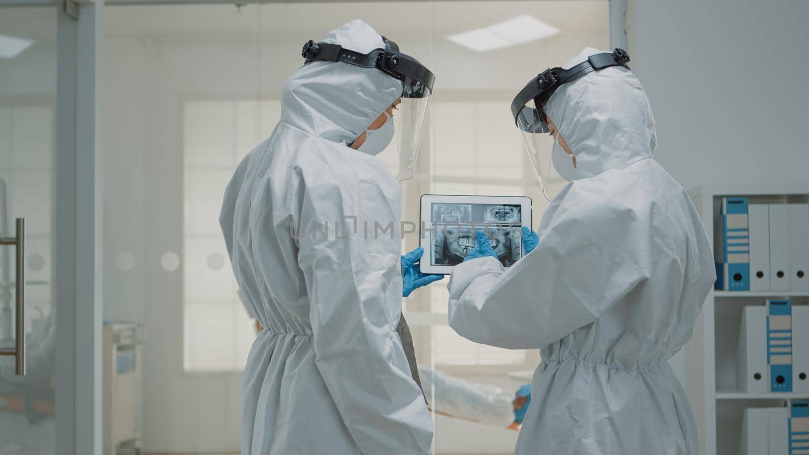 Professional orthodontists looking at dental x ray on digital tablet at clinic wearing protection suits during coronavirus epidemic. Dentists examining teeth radiography on scan device