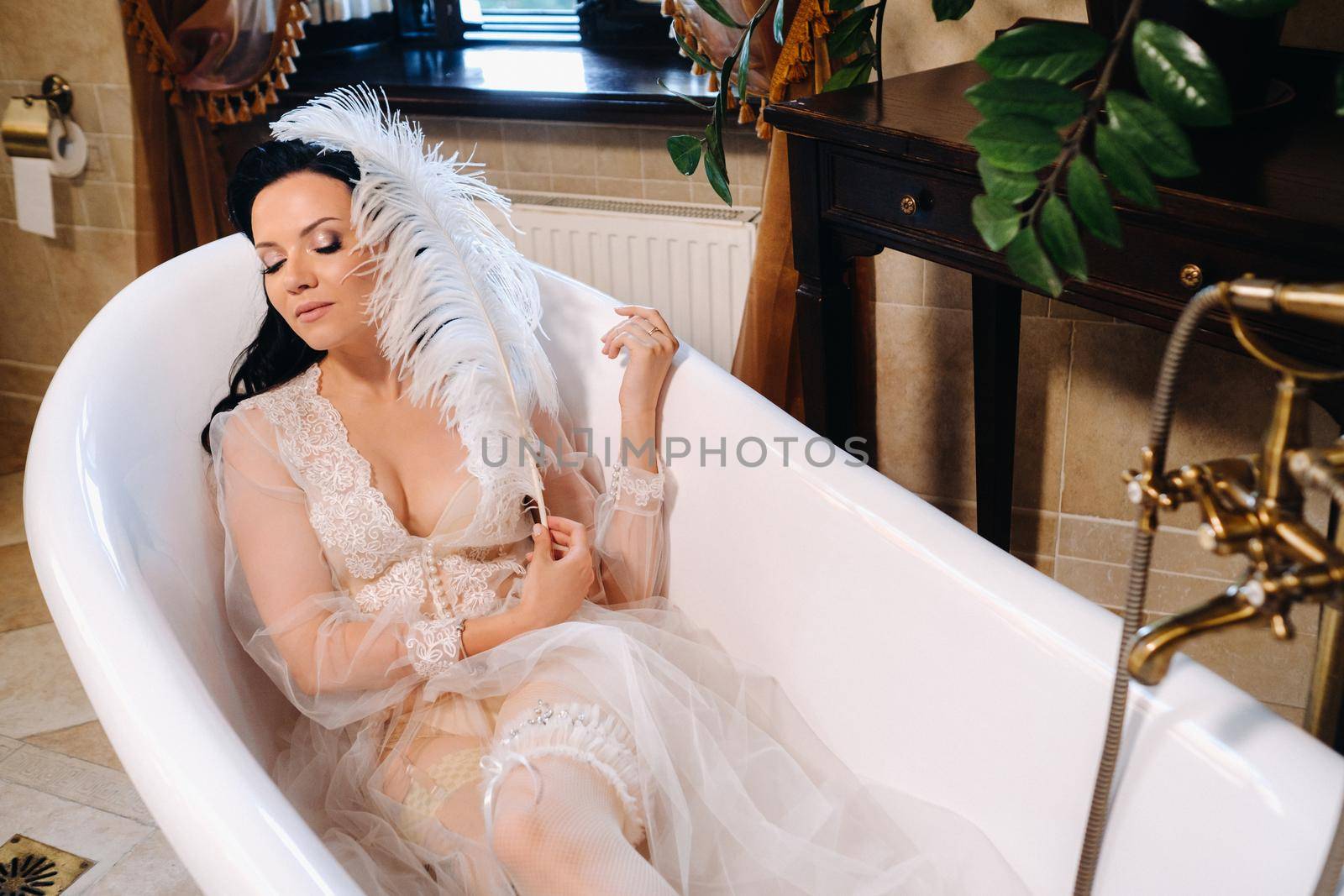 the bride, dressed in a boudoir transparent dress and underwear, lies in a vintage bathroom with a white feather in her hands, The morning of the bride.