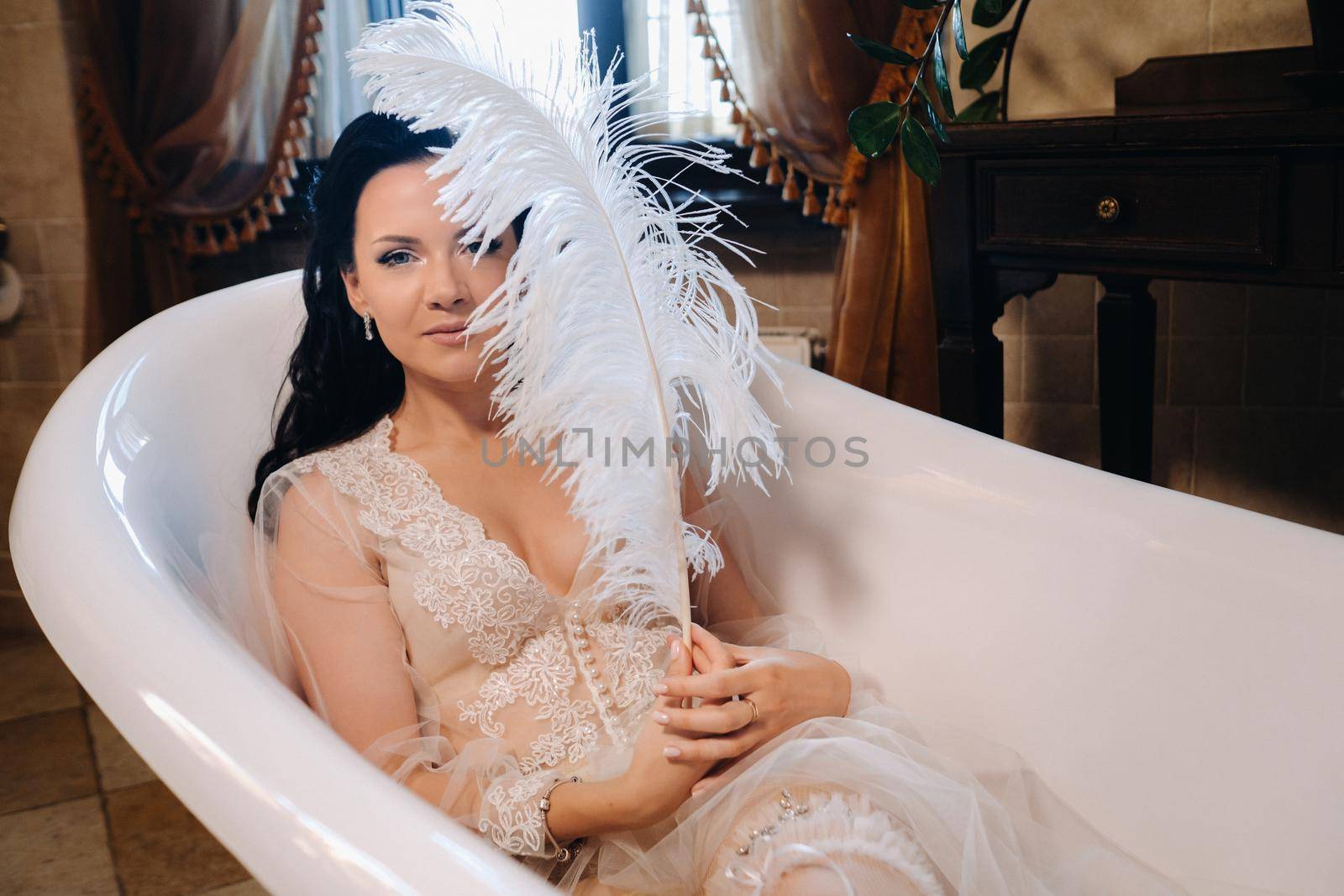 the bride, dressed in a boudoir transparent dress and underwear, lies in a vintage bathroom with a white feather in her hands, The morning of the bride.