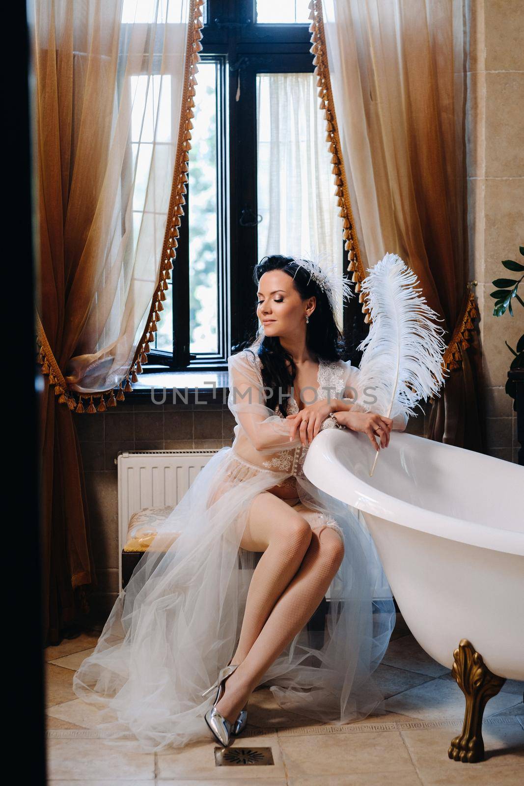 the bride, dressed in a boudoir transparent dress and underwear, sits near a vintage bath with a feather in her hands, The morning of the bride by Lobachad