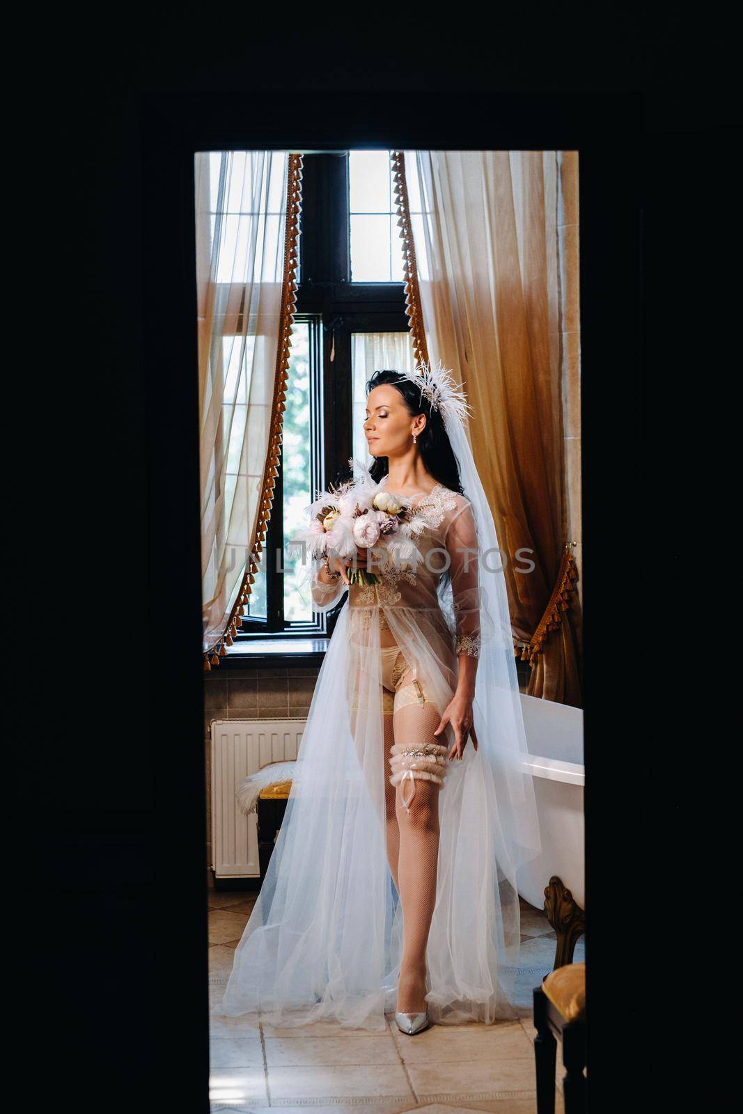 the bride, dressed in a boudoir transparent dress and underwear, sits near a vintage bath with a feather in her hands, The morning of the bride.