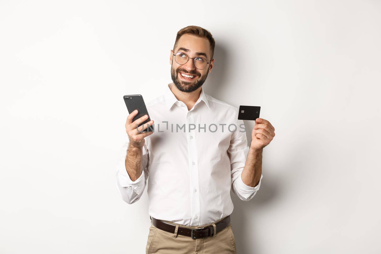 Business and online payment. Image of handsome man thinking while holding credit card and smartphone, standing against white background.