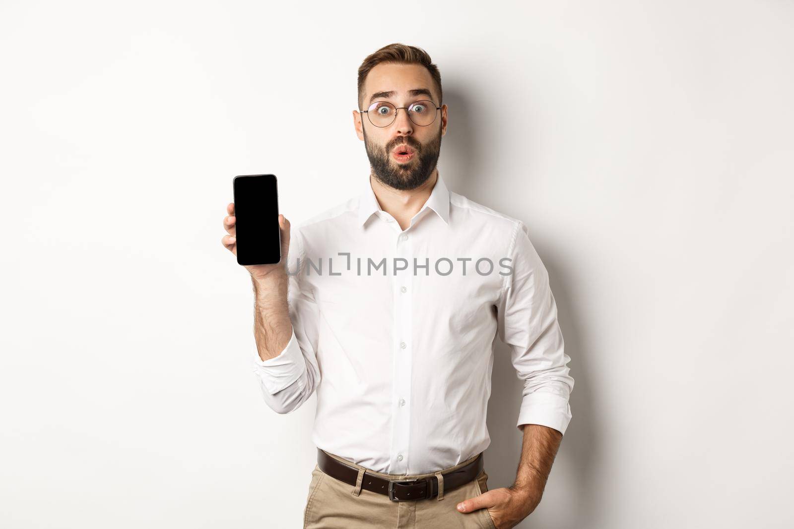 Surprised handsome manager in glasses, looking curious and showing mobile screen, standing over white background by Benzoix