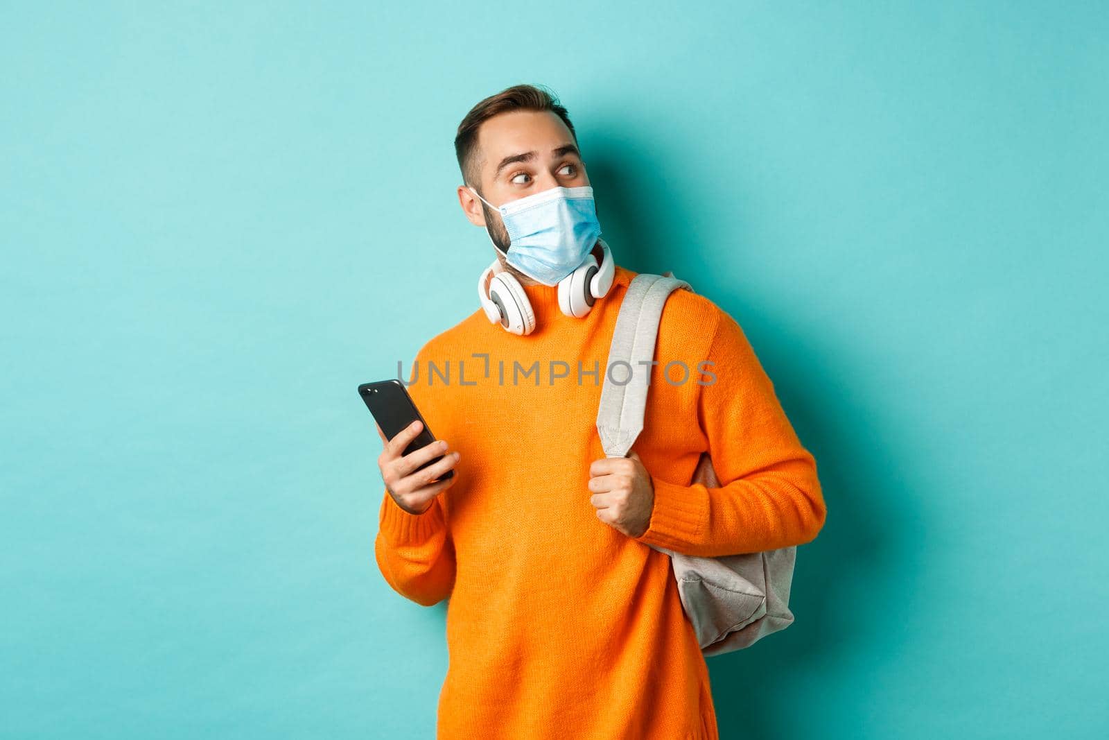 Young man in face mask using mobile phone, holding backpack, staring right amazed, standing against light blue background by Benzoix