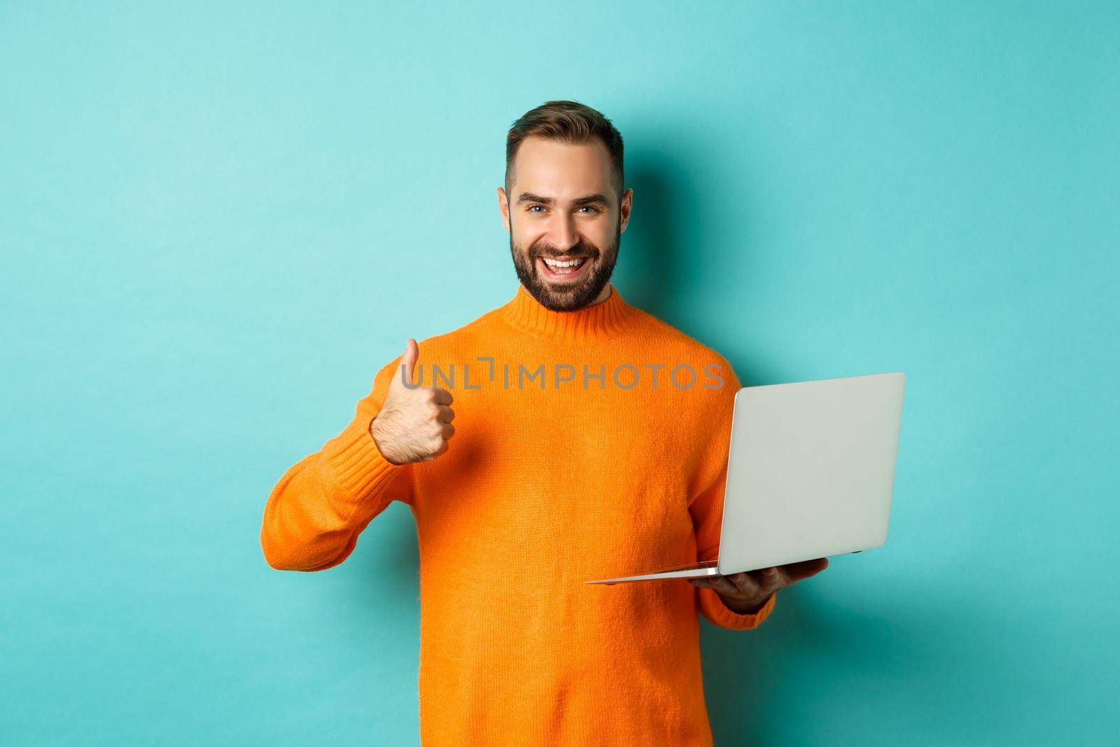 Freelance and technology concept. Lucky man in orange sweater, showing thumb up while working with laptop, standing satisfied over light blue background.