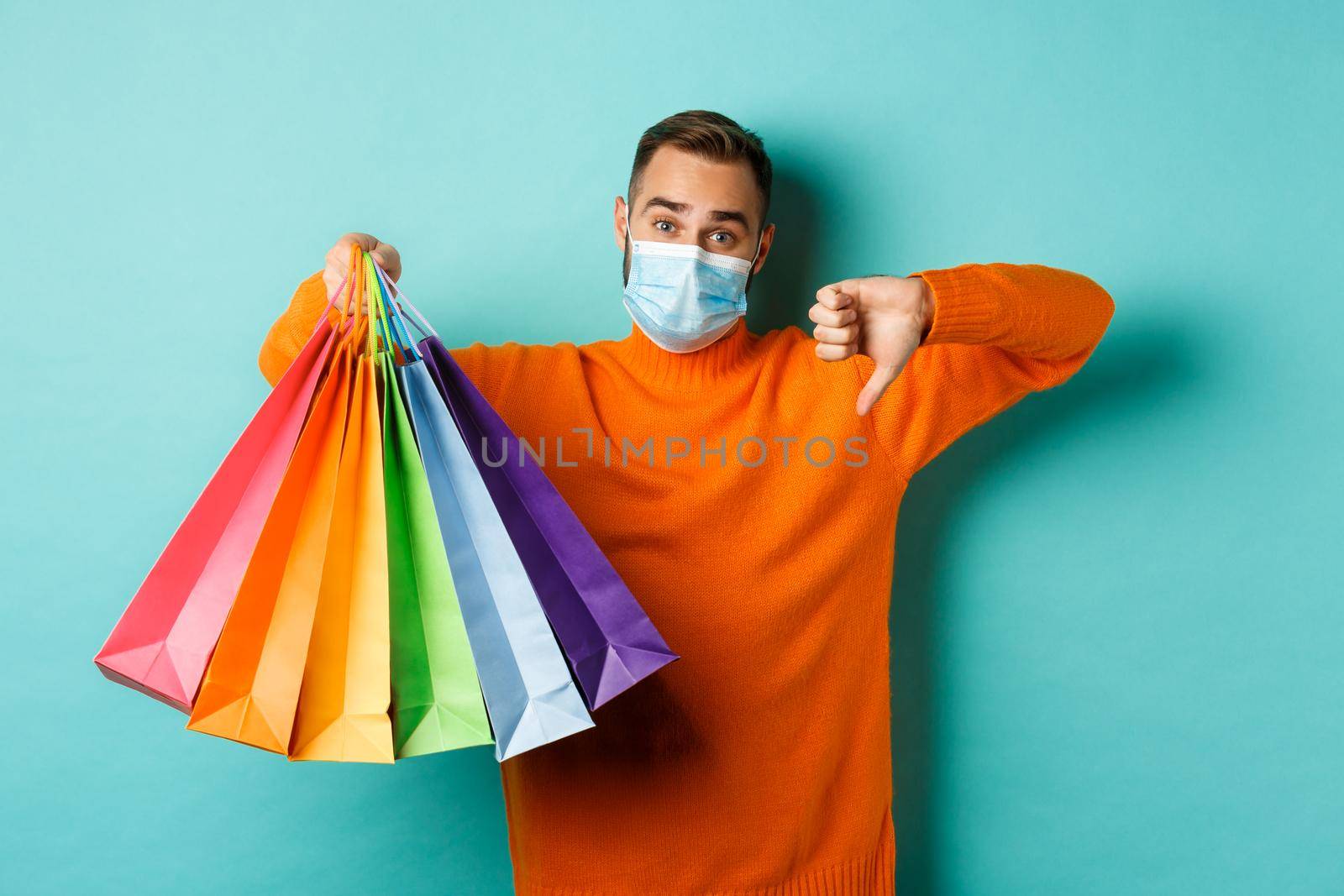 Covid-19, social distancing and lifestyle concept. Disappointed young man in medical mask from coronavirus, showing thumb down and shopping bags.