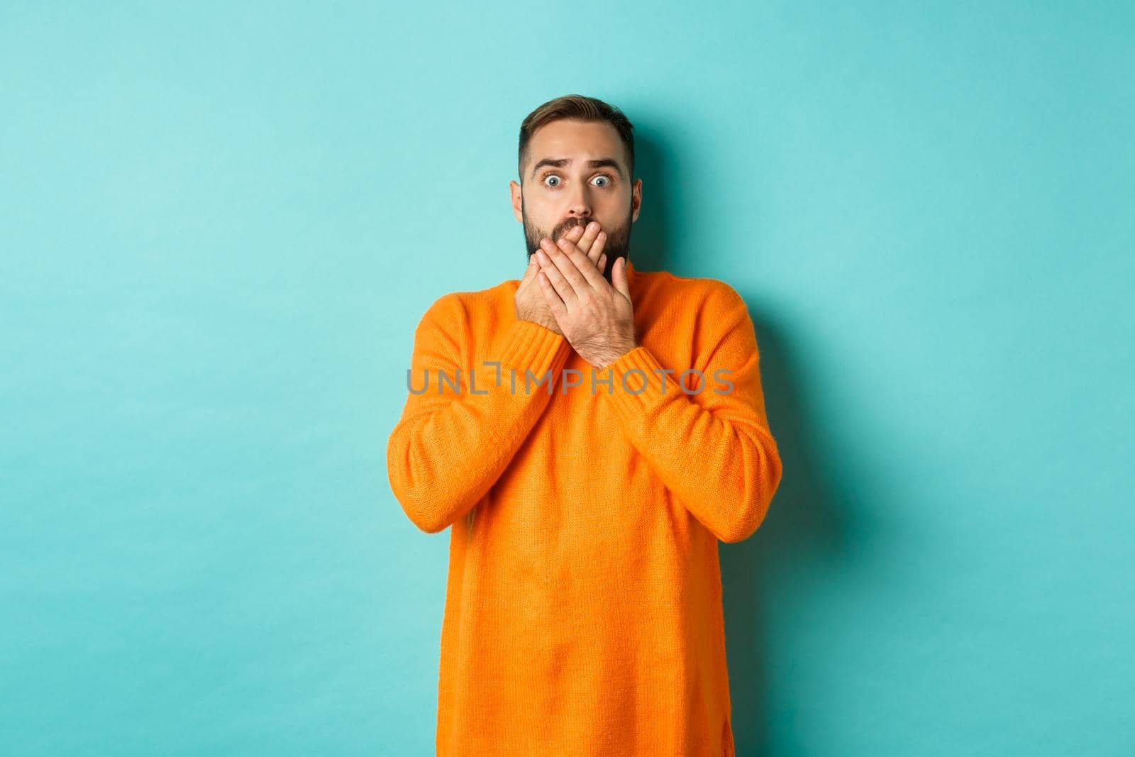 Startled man looking at horrible scene, gasping and stare scared, standing in orange winter sweater, studio background.