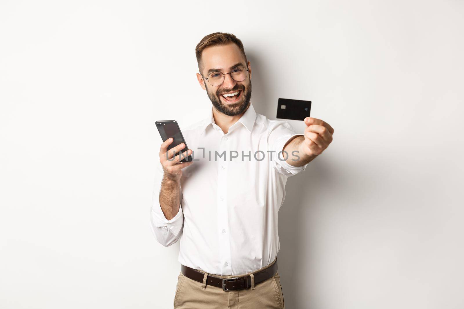 Business and online payment. Excited man showing his credit card while holding smartphone, standing satisfied against white background by Benzoix