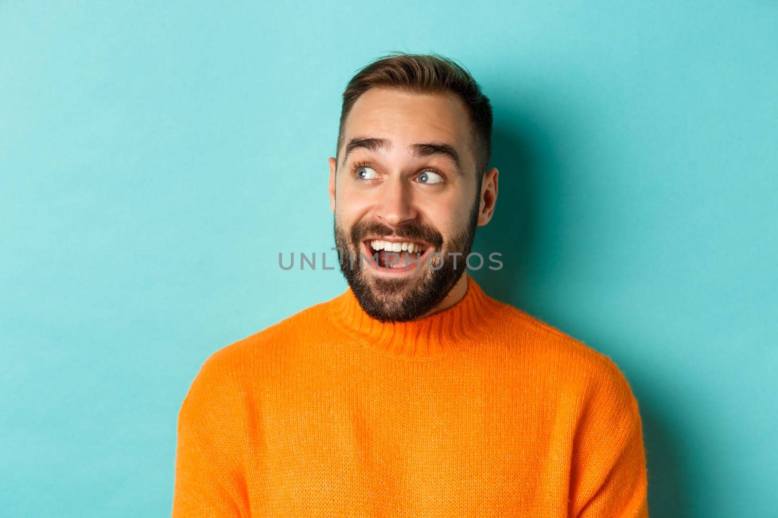 Close-up of handsome caucasian man smiling, looking left with surprised face, staring at logo, wearing orange sweater, standing against turquoise background.