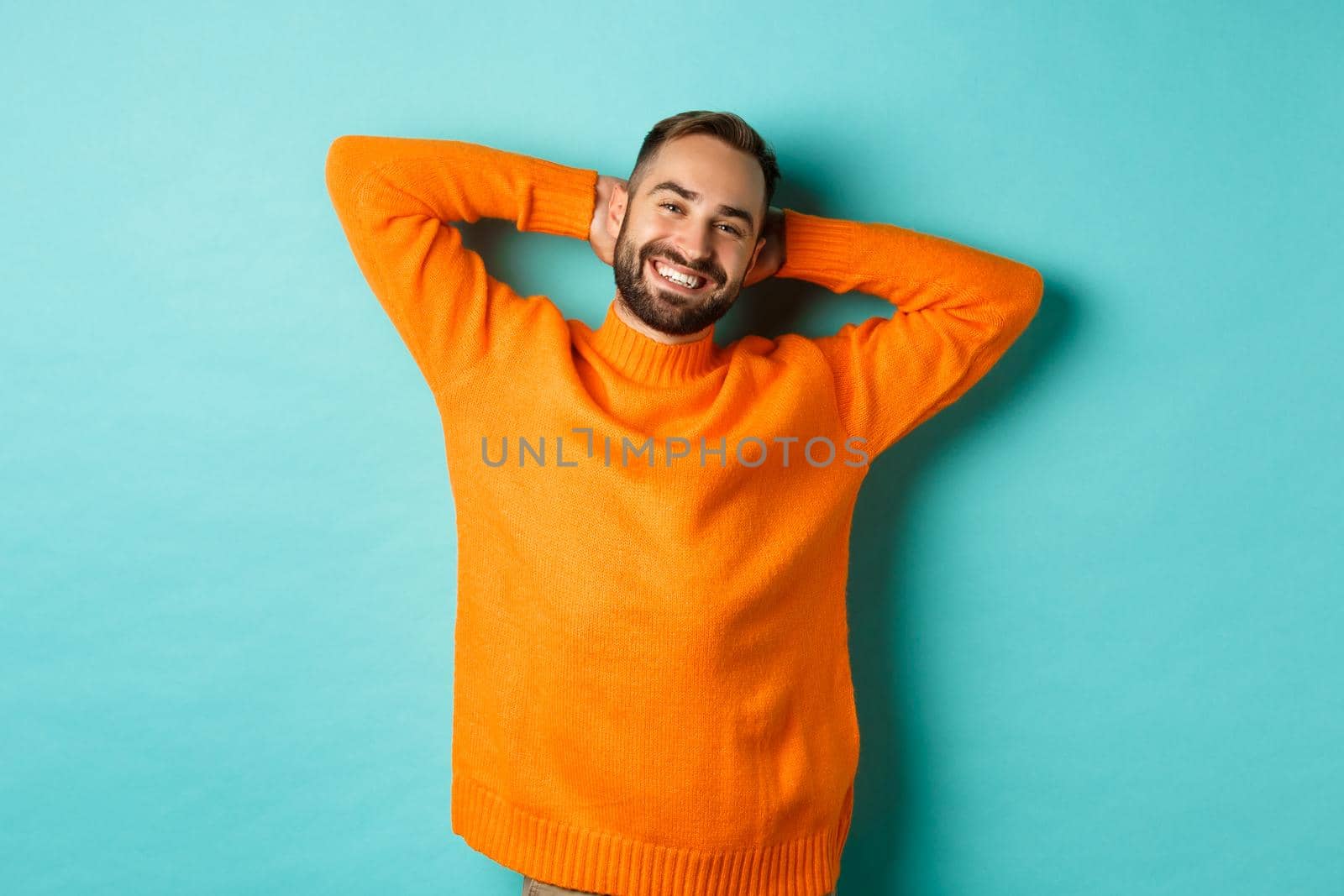 Handsome man relaxing, resting and smiling, holding hands behind head with carefree face, standing against light blue background.