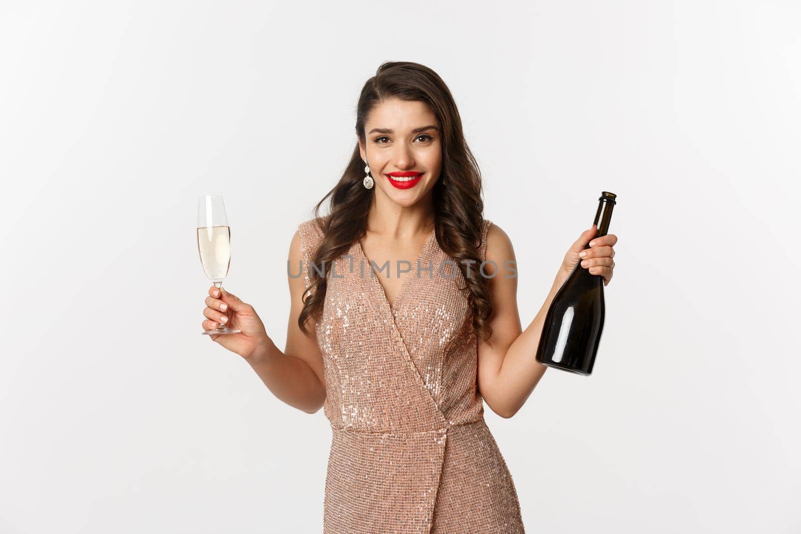 Winter holidays celebration concept. Elegant woman in luxury dress, drinking on New Year party, holding glass and bottle of champagne, standing over white background.