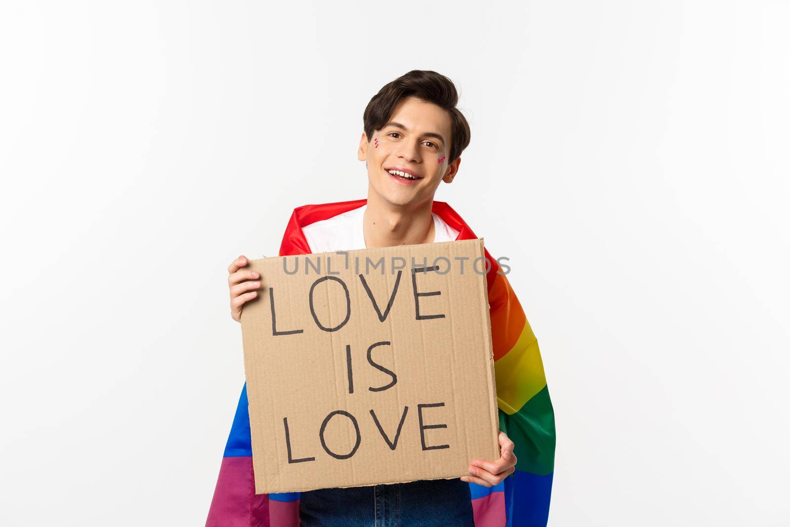 Smiling gay man activist holding sign love is love for lgbt pride parade, wearing Rainbow flag, standing over white background.