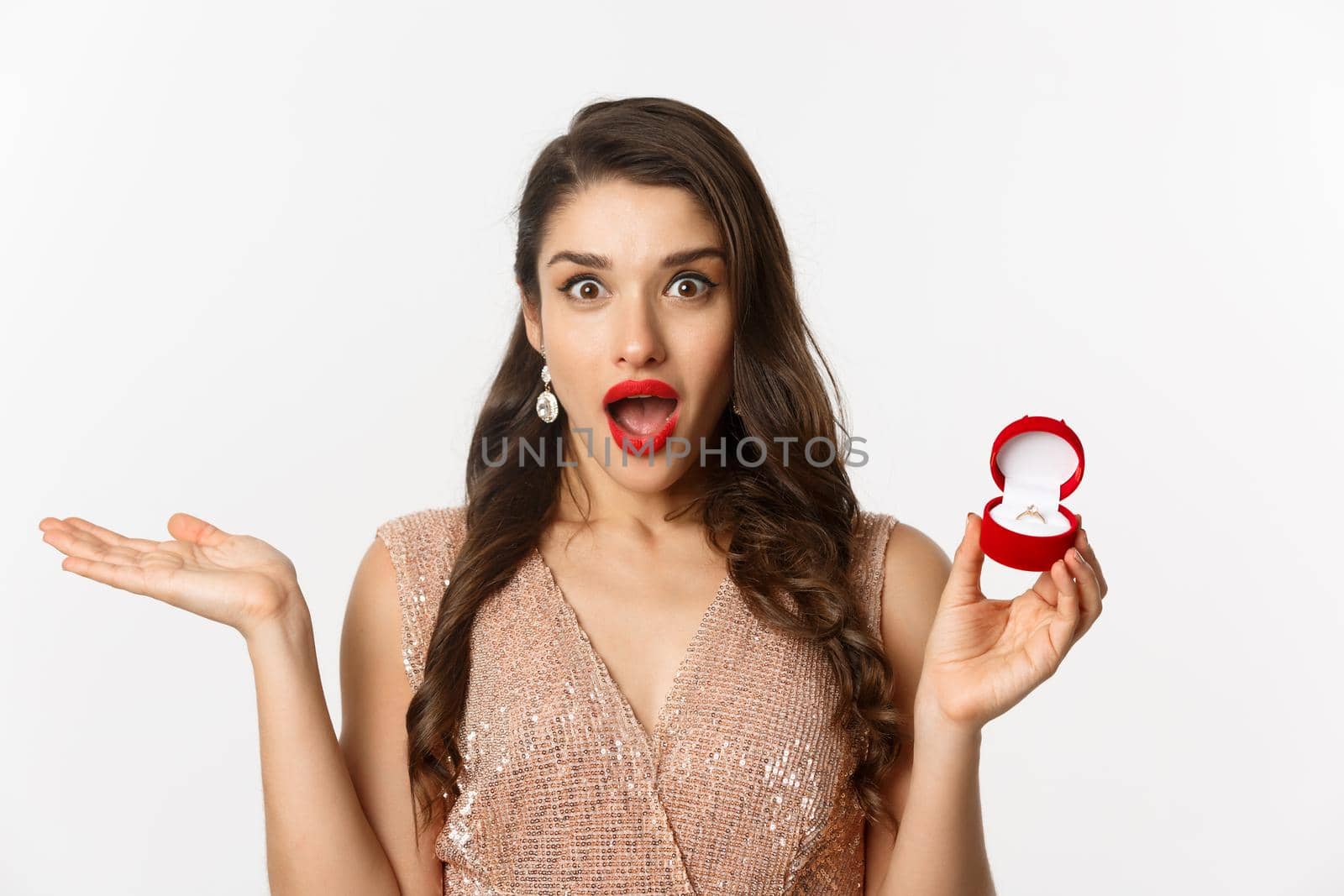 Close-up of attractive brunette woman with red lips, wearing dress, receiving engagement ring and looking surprise, standing over white background.