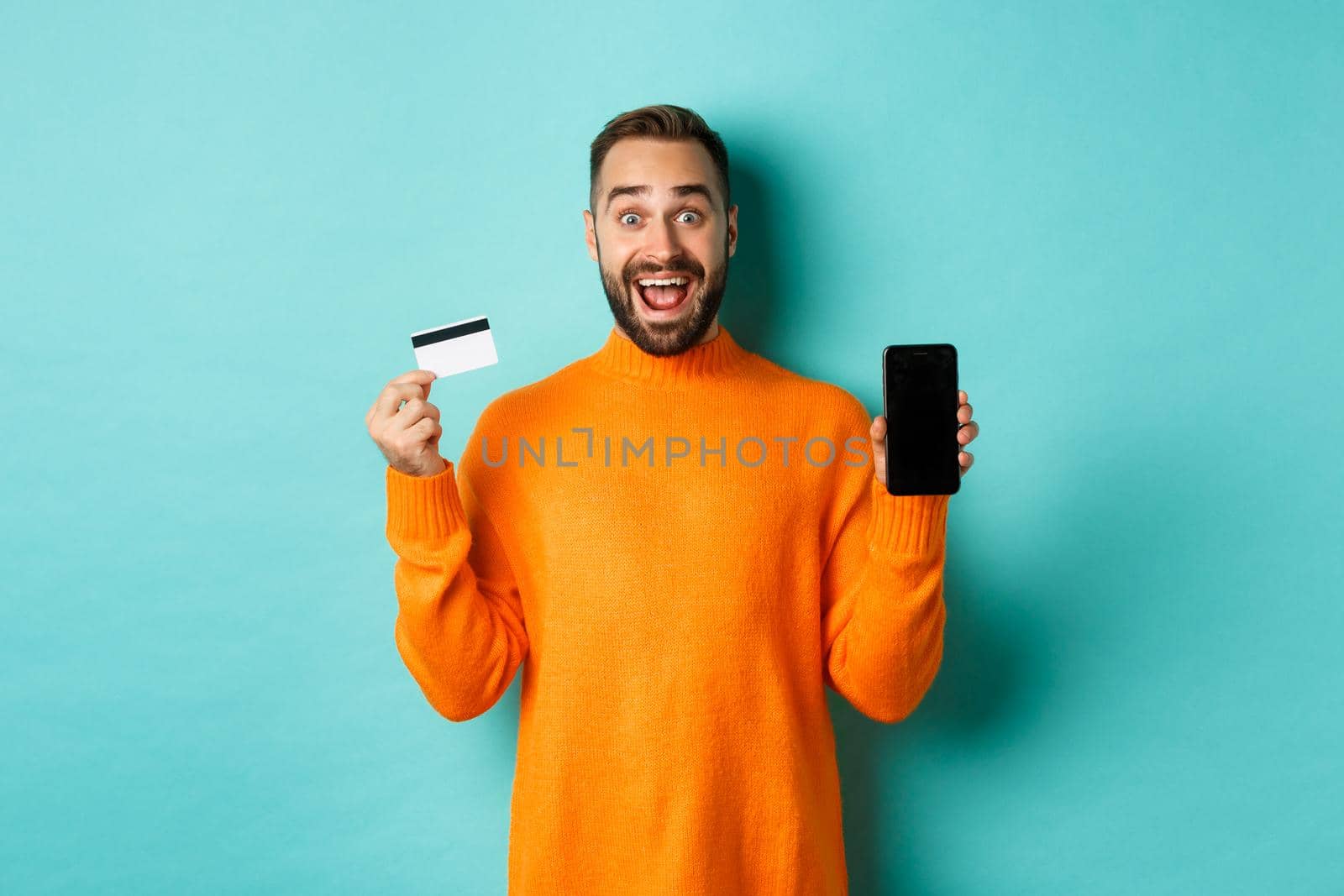 Online shopping. Excited man showing mobile screen and credit card, smiling amazed, standing over turquoise background.