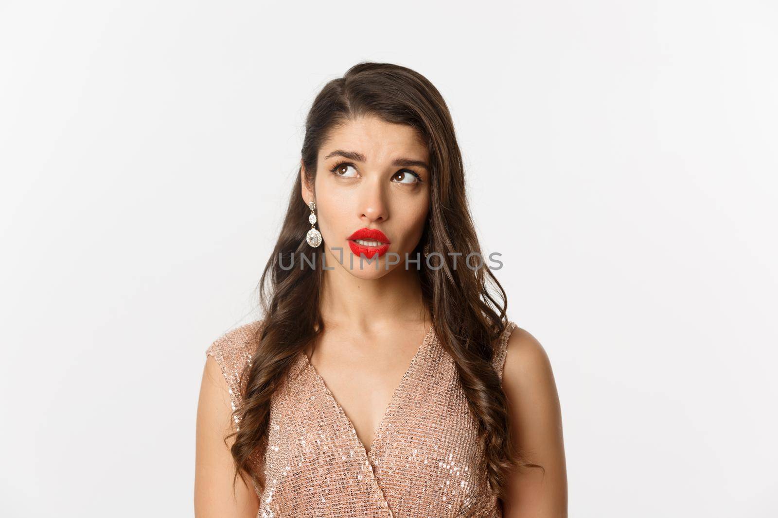 Concept of New Year celebration and winter holidays. Close-up of elegant woman with red lips and dress, looking at upper left corner and thinking, standing over white background.