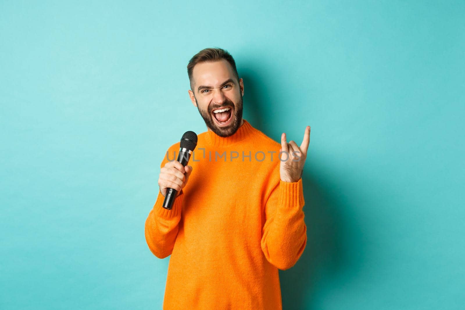 Joyful handsome man singing song in microphone, showing rock-n-roll gesture excited, standing over turquoise background.