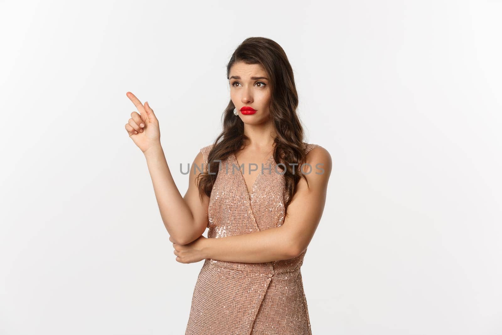 Christmas, holidays and celebration concept. Skeptical and displeased woman in stylish party dress, pointing finger left and starring at banner unamused, white background.