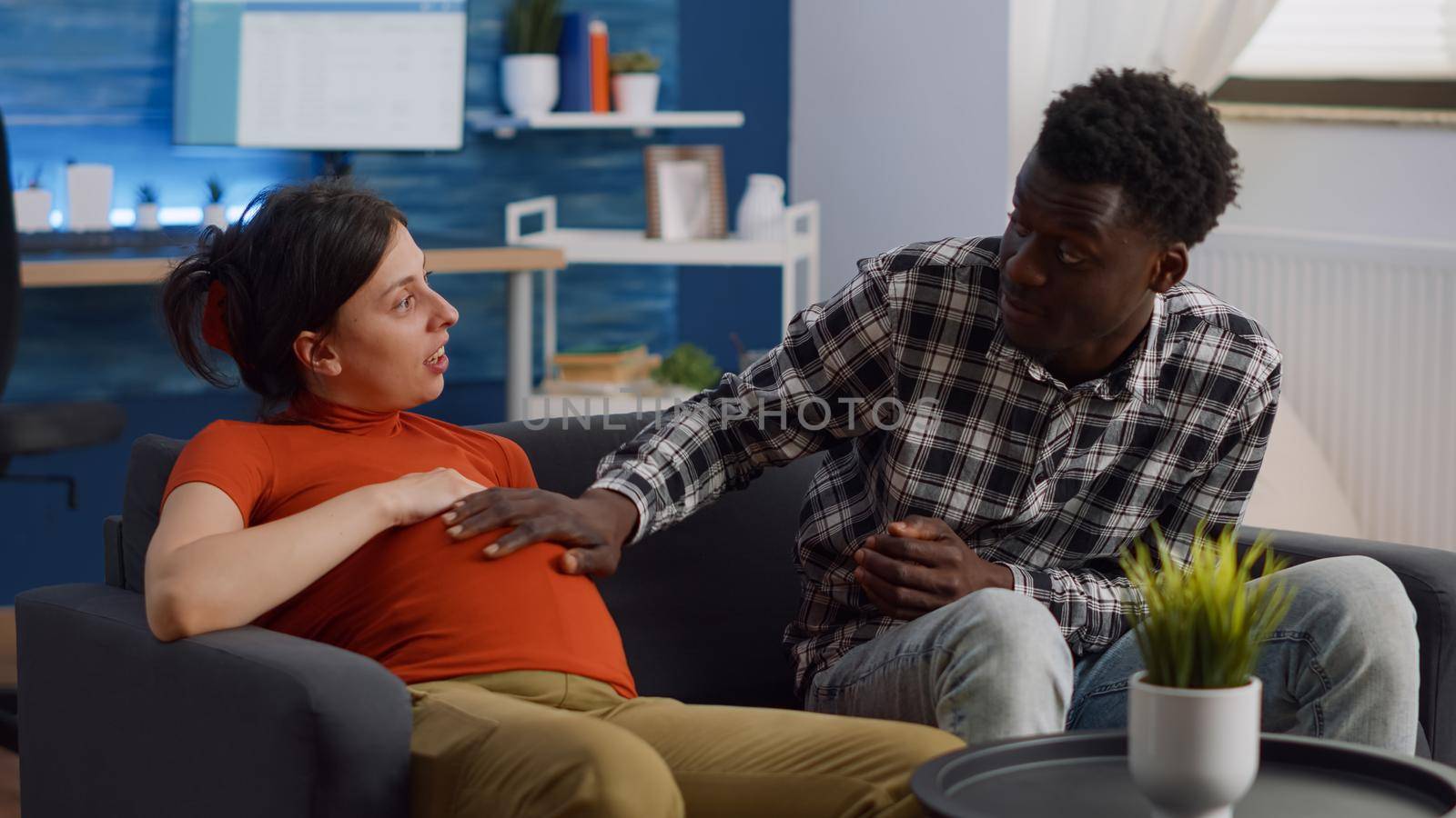 Young interracial couple with pregnancy chatting about baby at home. African american father of child touching belly while pregnant caucasian woman relaxing on sofa in living room.