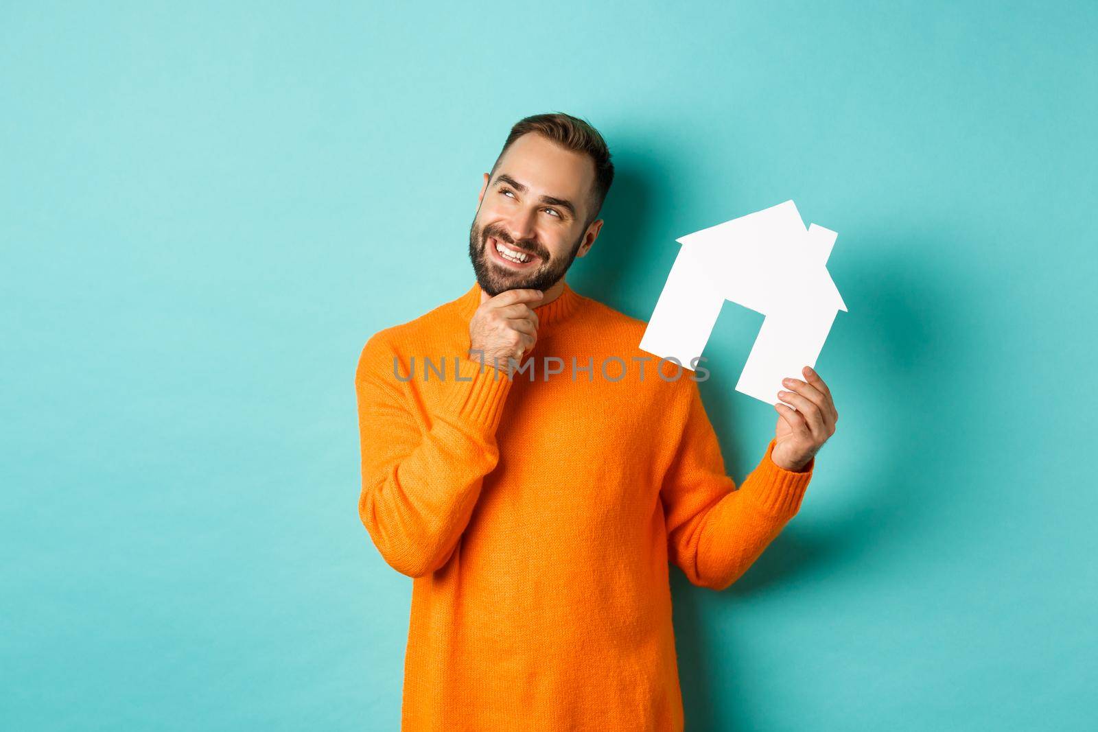 Real estate concept. Handsome adult man in orange sweater thinking about buying flat or renting, holding house maket and looking thoughtful, standing over blue background.