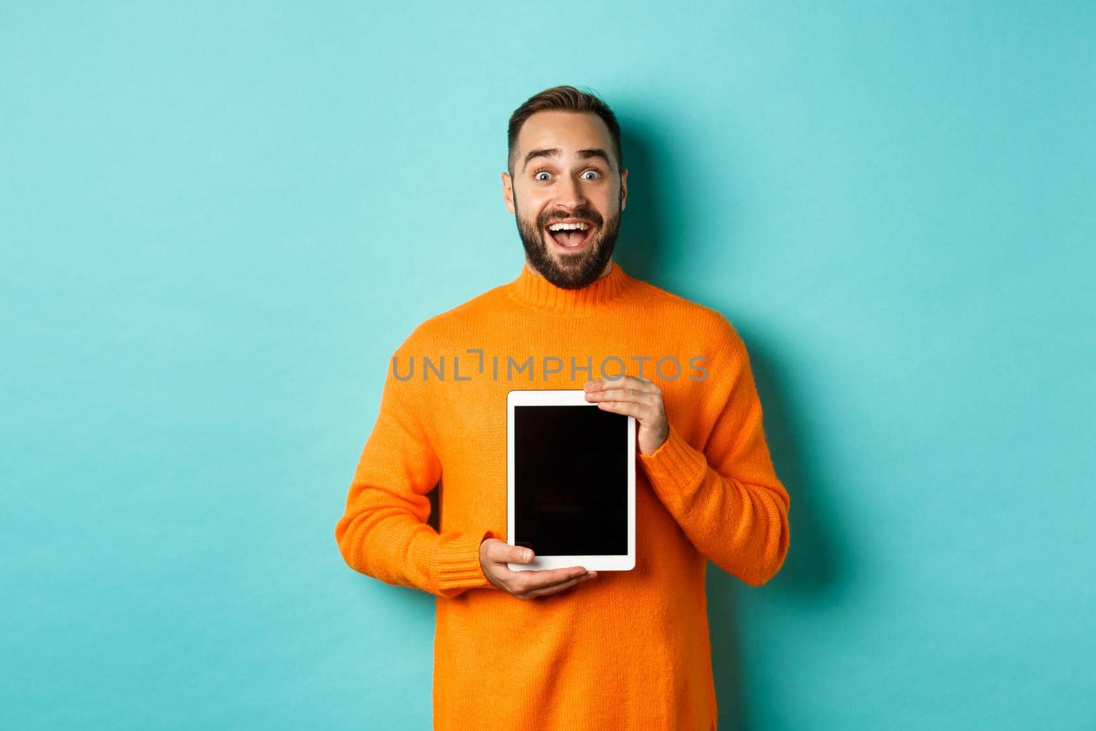 Technology. Excited adult man in orange sweater showing digital tablet screen, staring amazed at camera, standing over turquoise background.