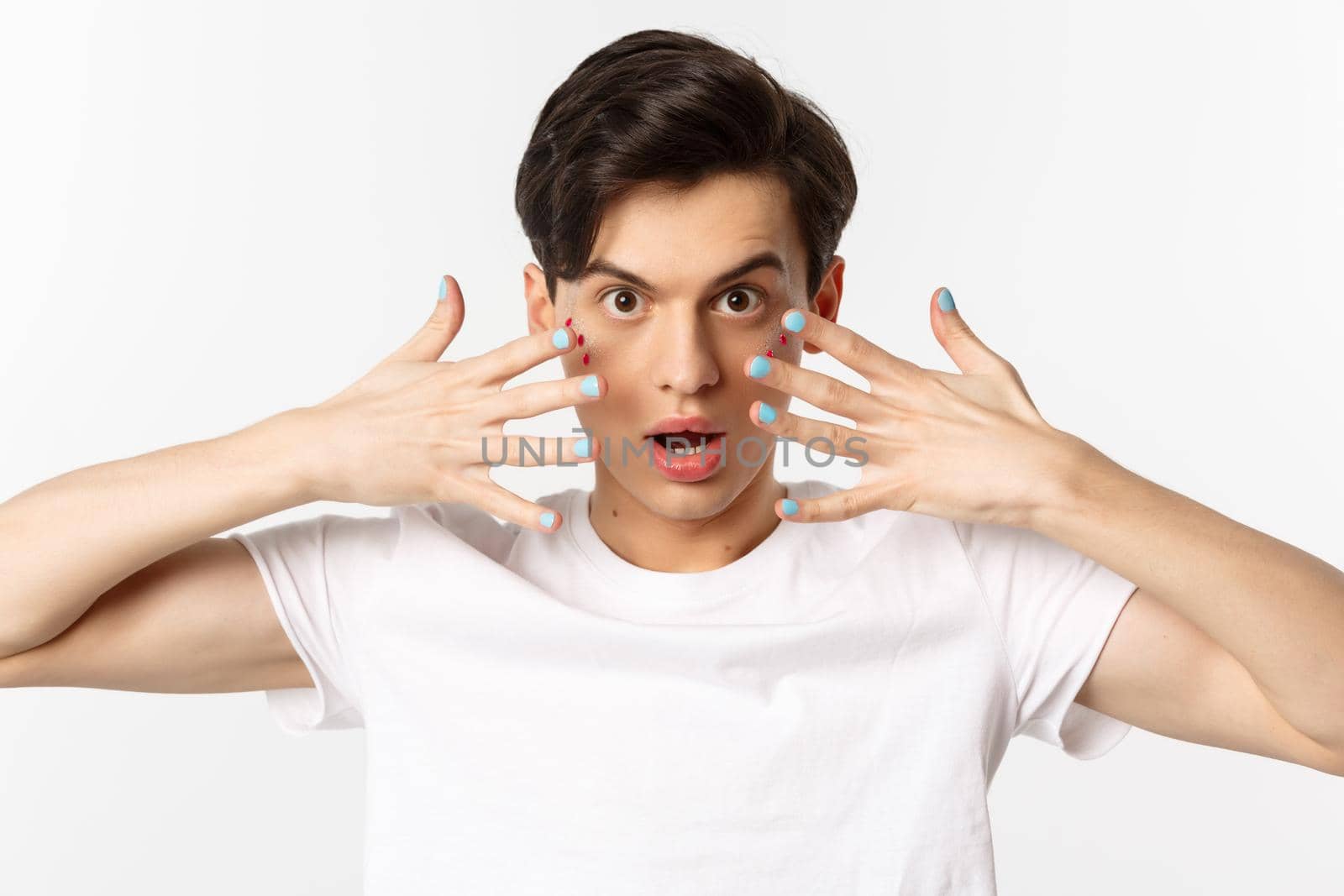 People, lgbtq and beauty concept. Close-up of beautiful gay man with glitter on face, showing nail polish on his fingernails, looking sassy at camera, standing over white background.