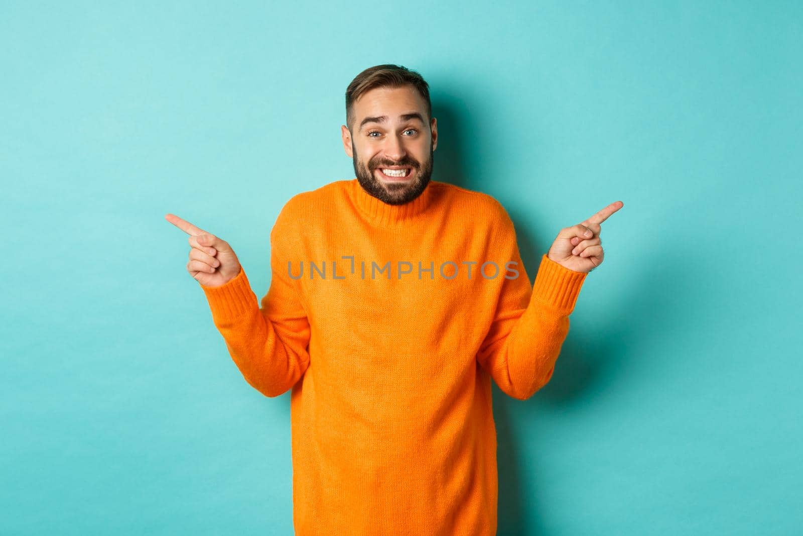 Confused handsome man shrugging indecisive, pointing fingers sideways, cant choose between two variants, standing against light blue background.