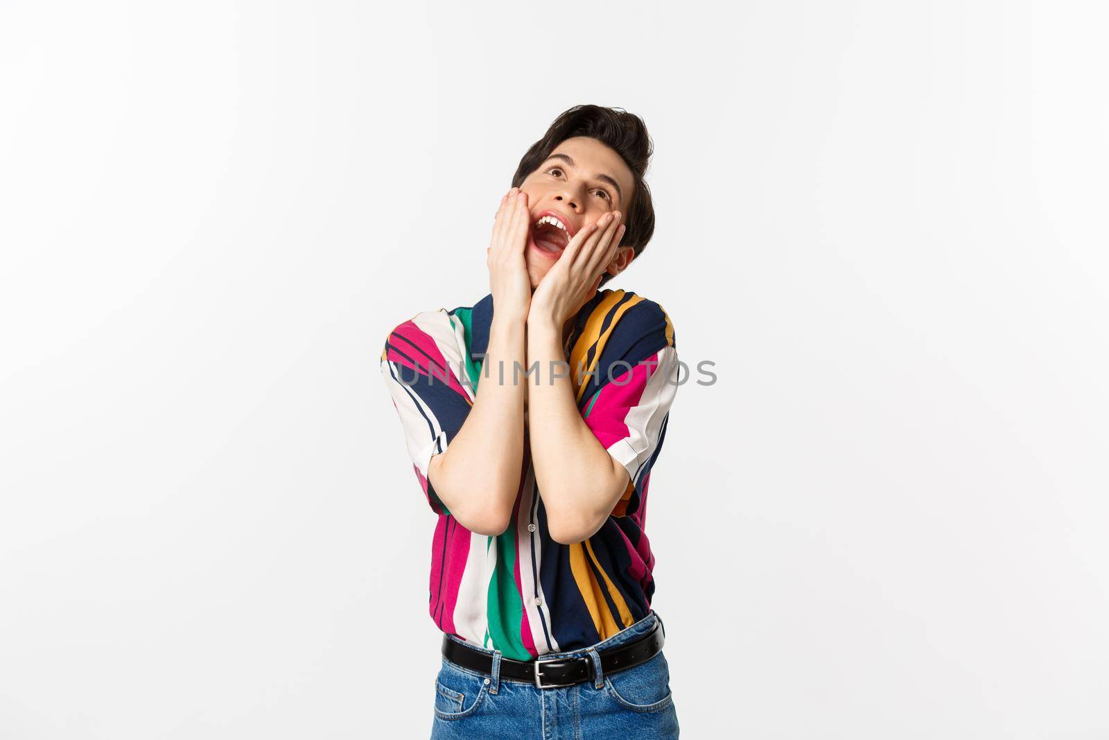 Image of excited gay man shouting of joy and looking up, feeling ecstatic, standing against white background.