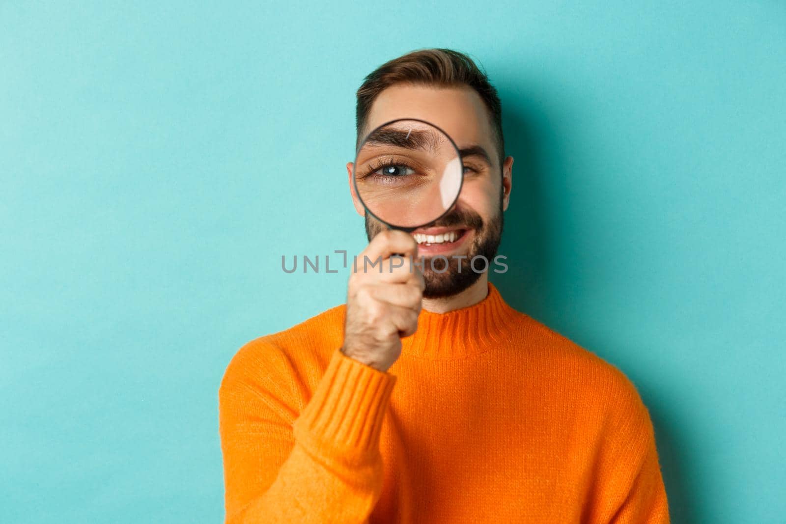 Funny man looking through magnifying glass, searching or investigating something, standing in orange sweater against turquoise background by Benzoix