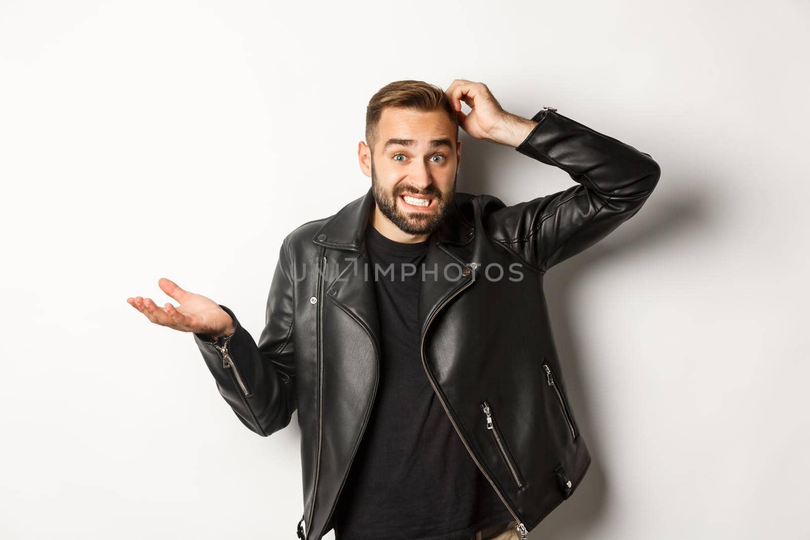 Confused bearded guy in cool leather jacket shrugging, scratch head puzzled and clueless, standing over white background.