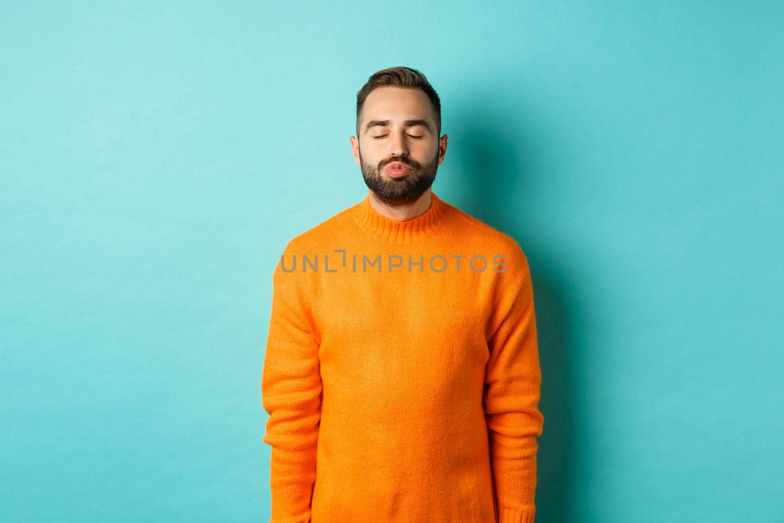 Waist up shot of young man pucker lips and close eyes, waiting for kiss, standing in orange sweater against light blue background.
