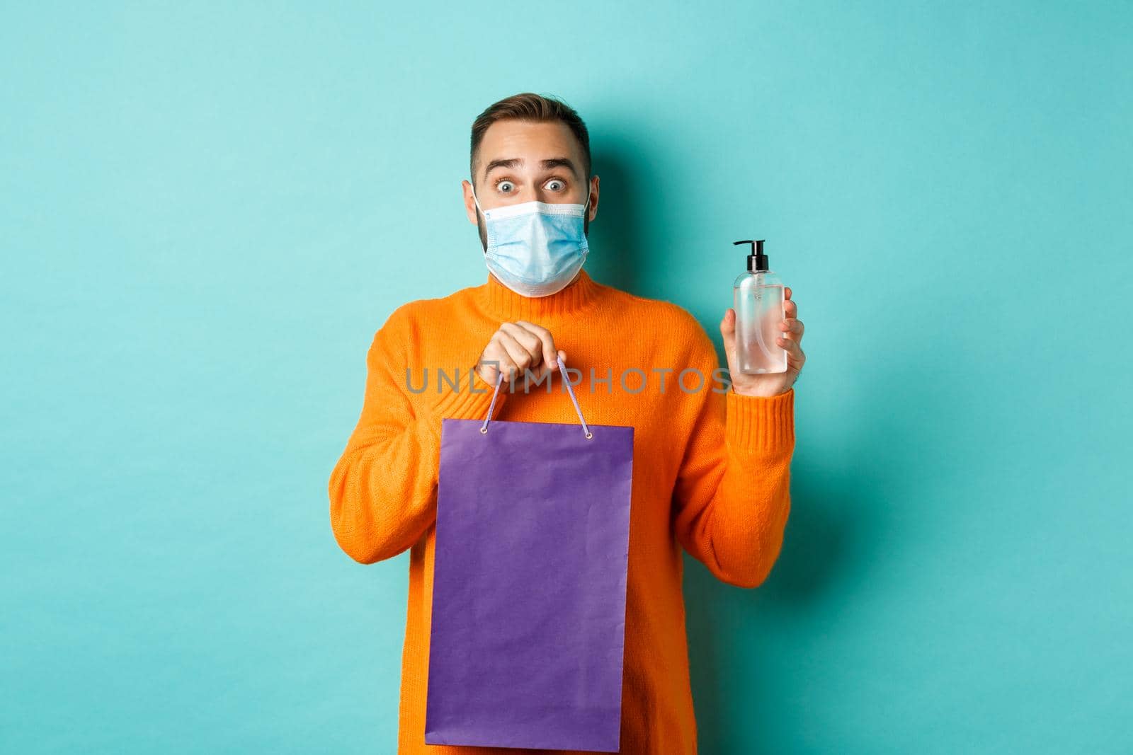 Coronavirus, pandemic and lifestyle concept. Man in face mask showing shopping bag and hand sanitizer, standing over turquoise background by Benzoix
