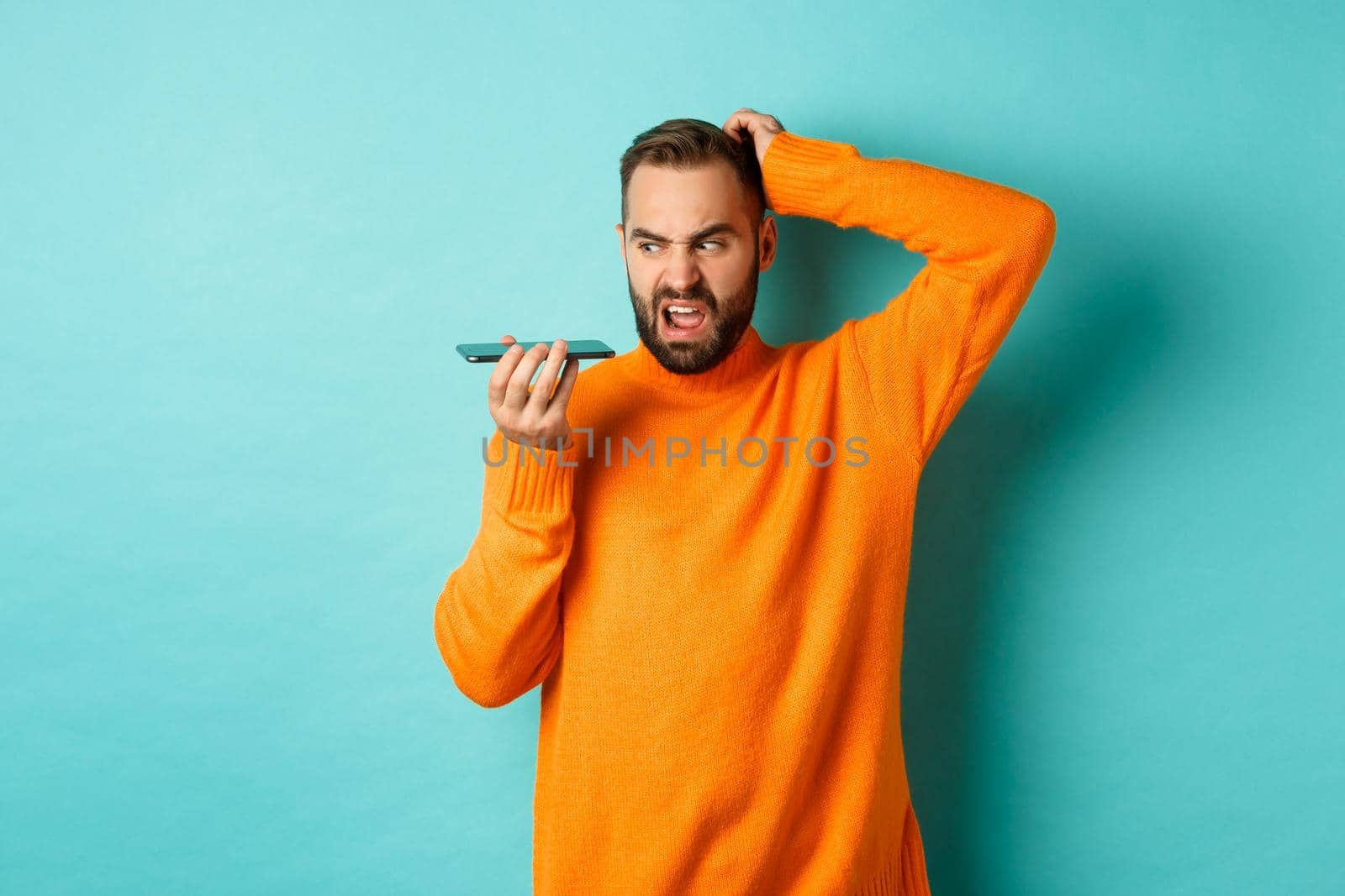 Confused man scratching head while talking on speakerphone, record voice message with indecisive face, standing in orange sweater over light blue background.
