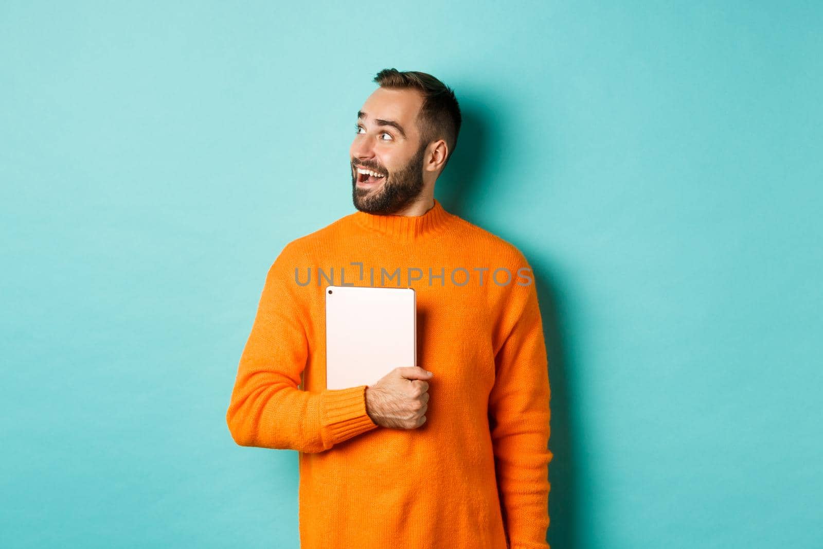 Handsome man holding laptop, looking left with surprise and amazement, standing in orange sweater against light blue background by Benzoix