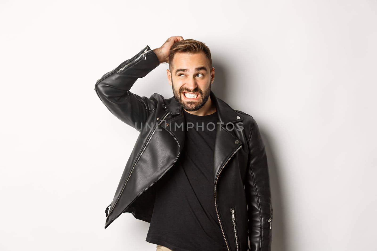 Confused macho guy in black leather jacket looking awkward and unsure, scratching head puzzled and stare left, white background.