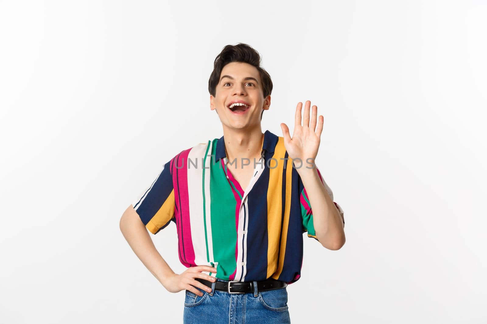 Cheerful gay man saying hello, waving hand and greeting person, standing over white background.