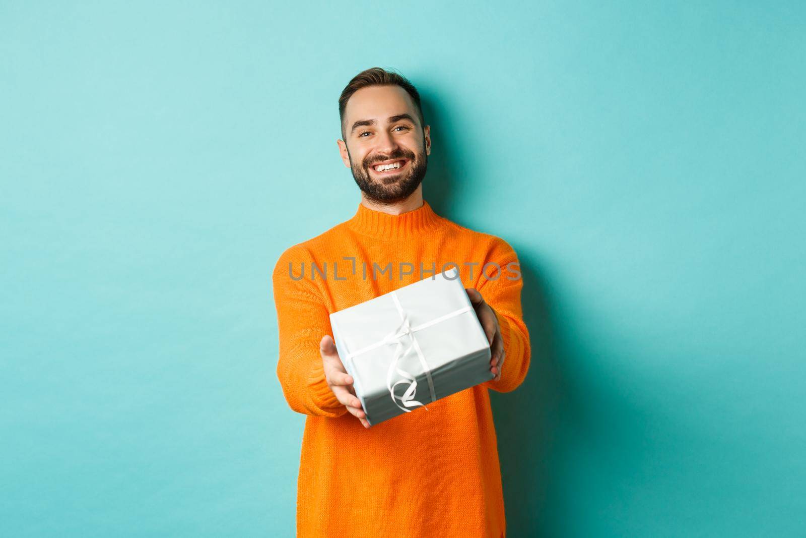 Holidays and celebration concept. Happy man giving you a gift, extending hand with present and smiling, standing over turquoise background.