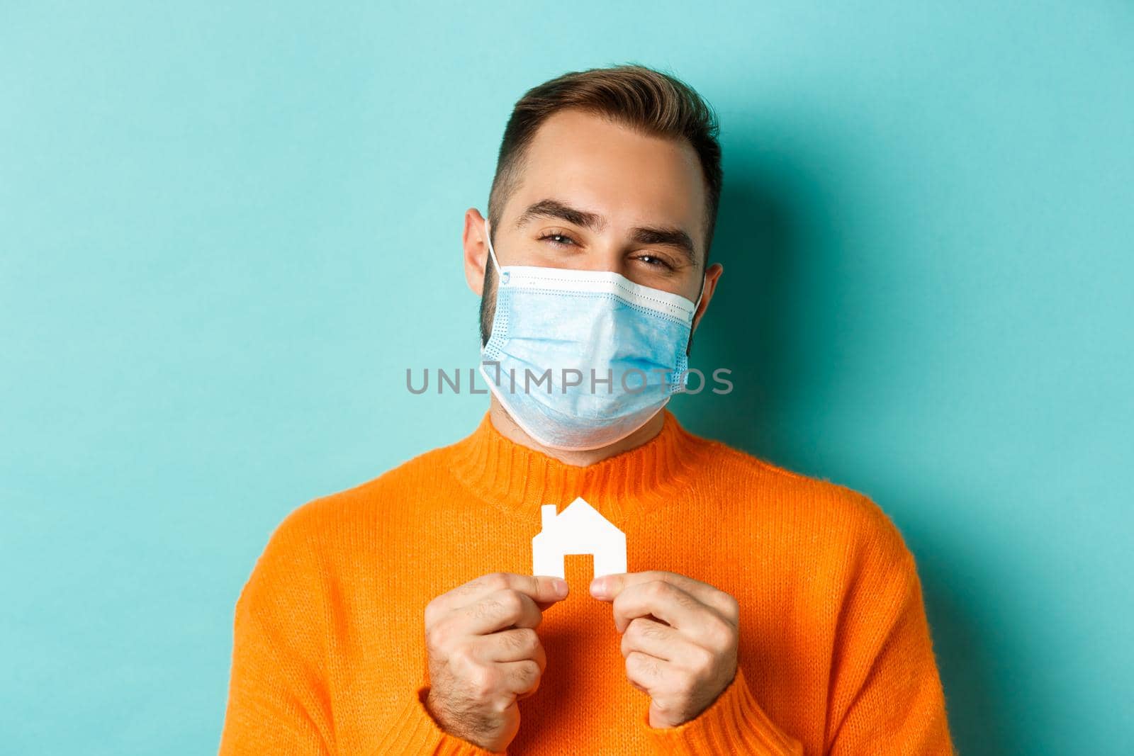 Real estate and coronavirus pandemic concept. Close-up of adult man in medical mask holding small paper house maket and smiling, searching for apartment, light blue background by Benzoix