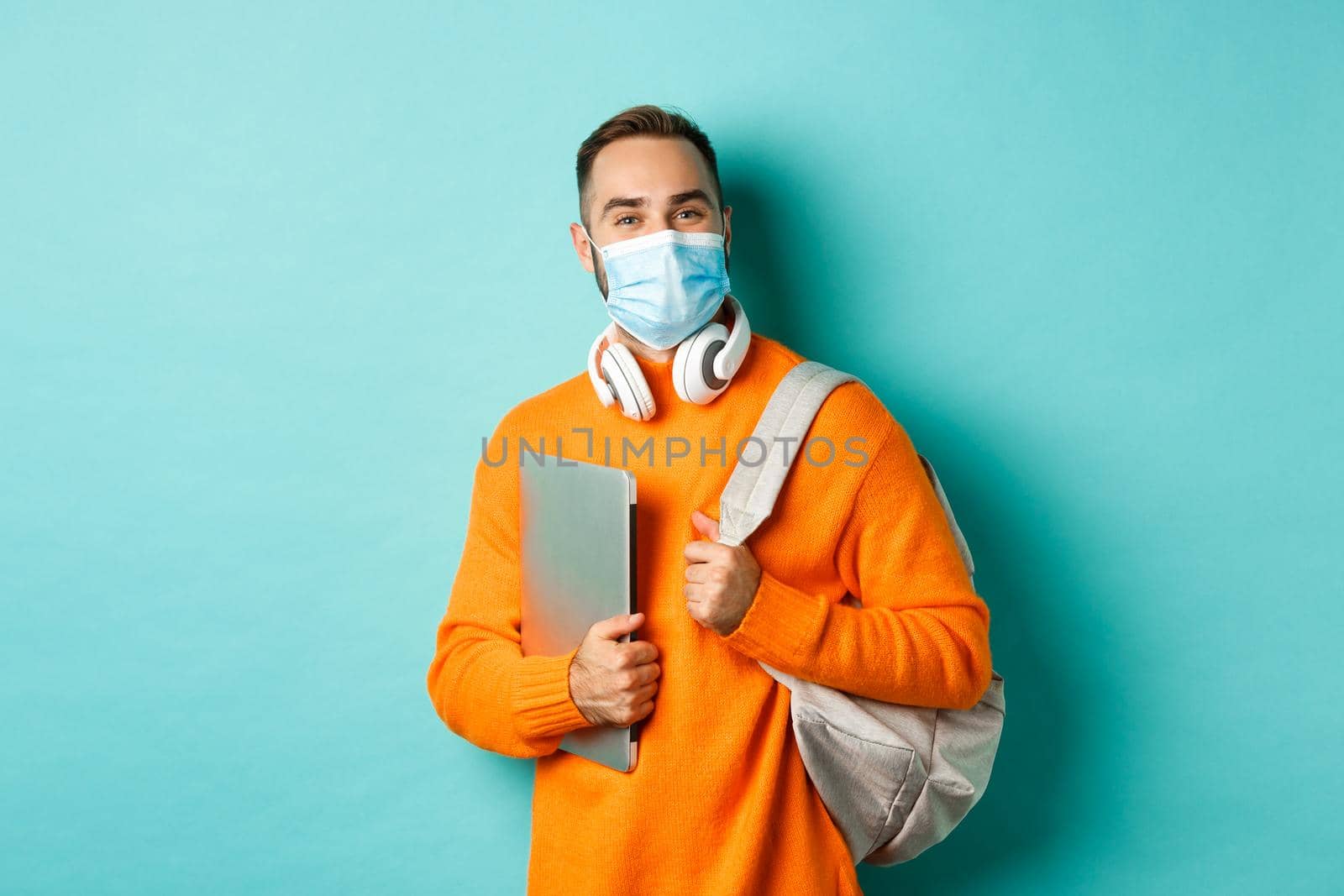 Handsome student in face mask, holding backpack and laptop, going work, standing over light blue background.